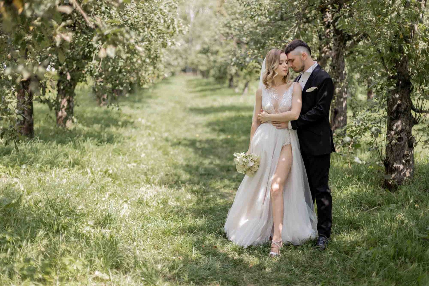 Bride and groom embrace romantically in a lush green orchard, bride wearing elegant white gown with slit holding bouquet.