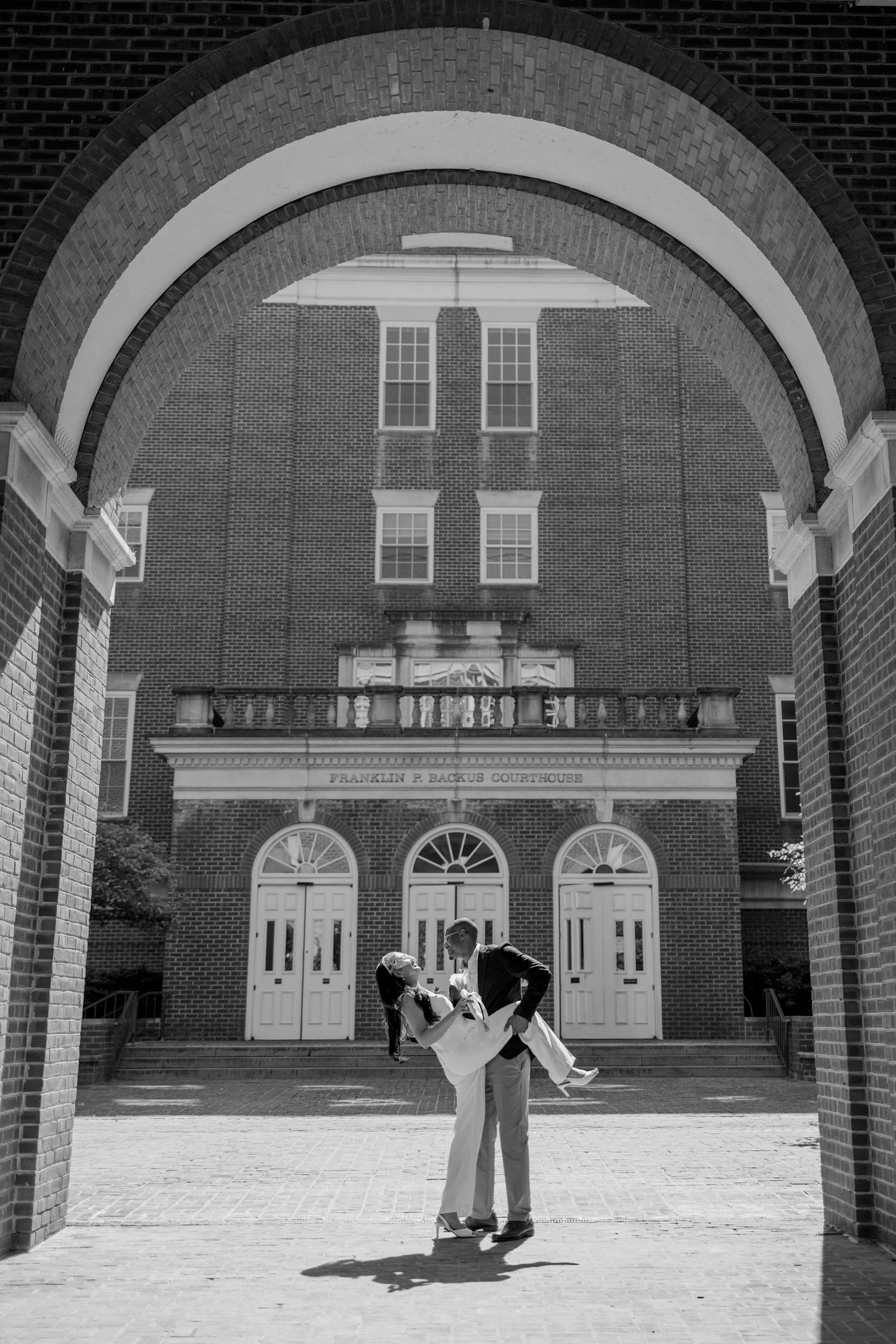 Black and white photo of couple embracing under brick archway outside elegant building.