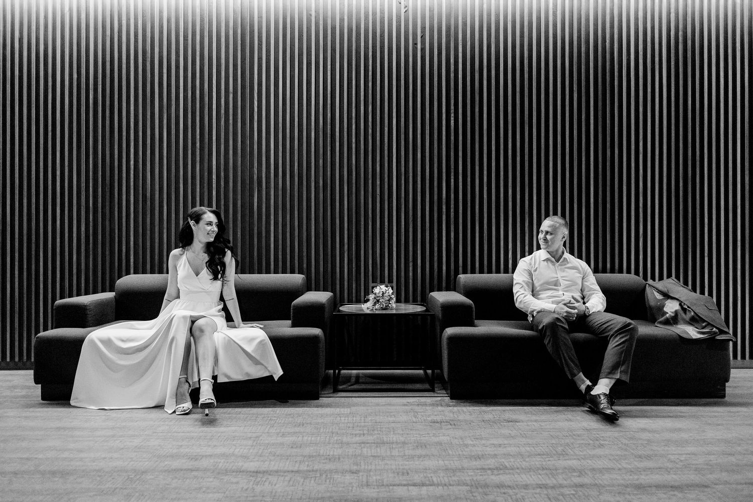 Black and white photo of a bride and groom sitting apart on modern sofas against a striped wall.