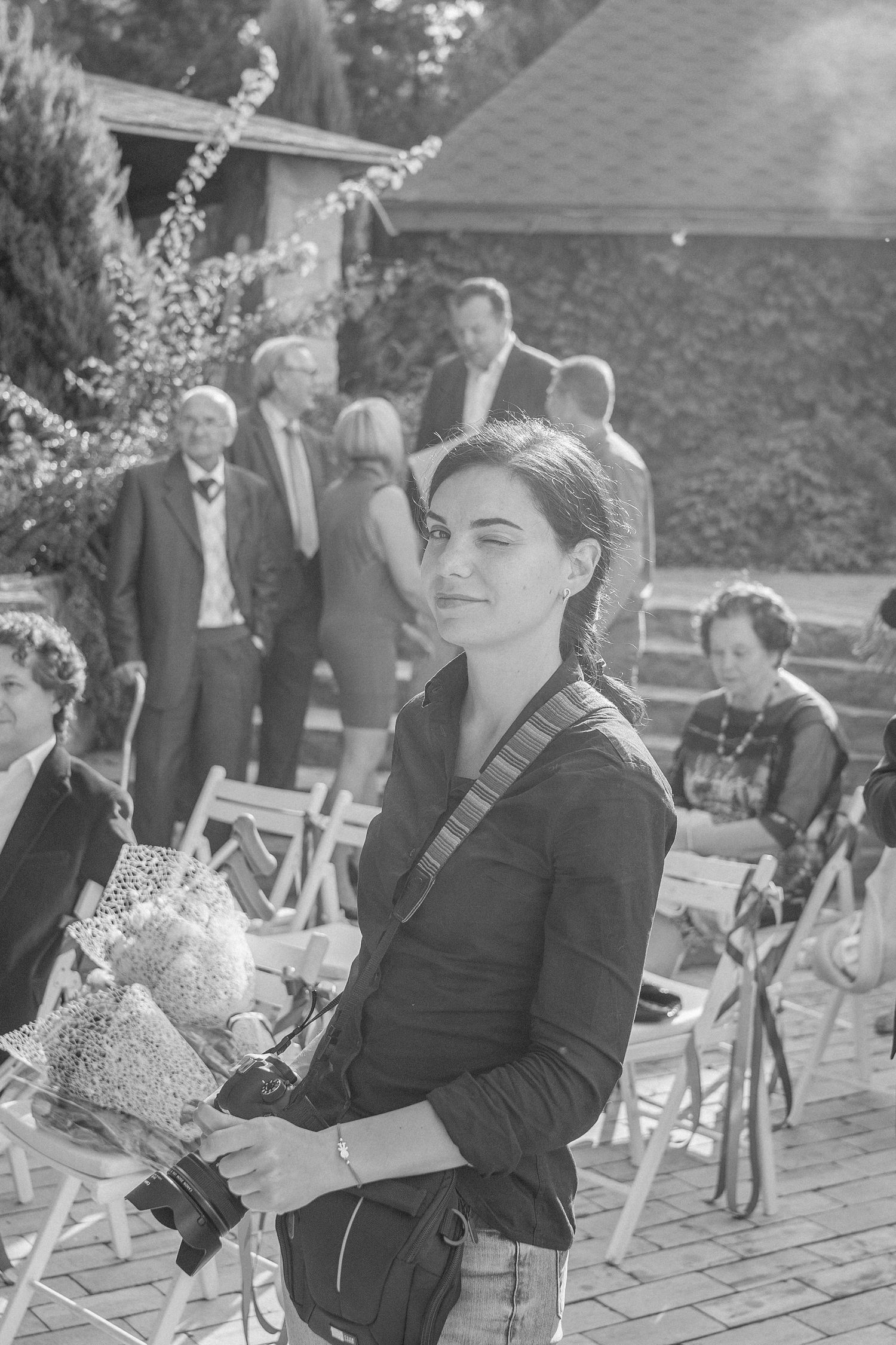 Black and white photo of a young woman holding flowers and a camera at an outdoor wedding ceremony.