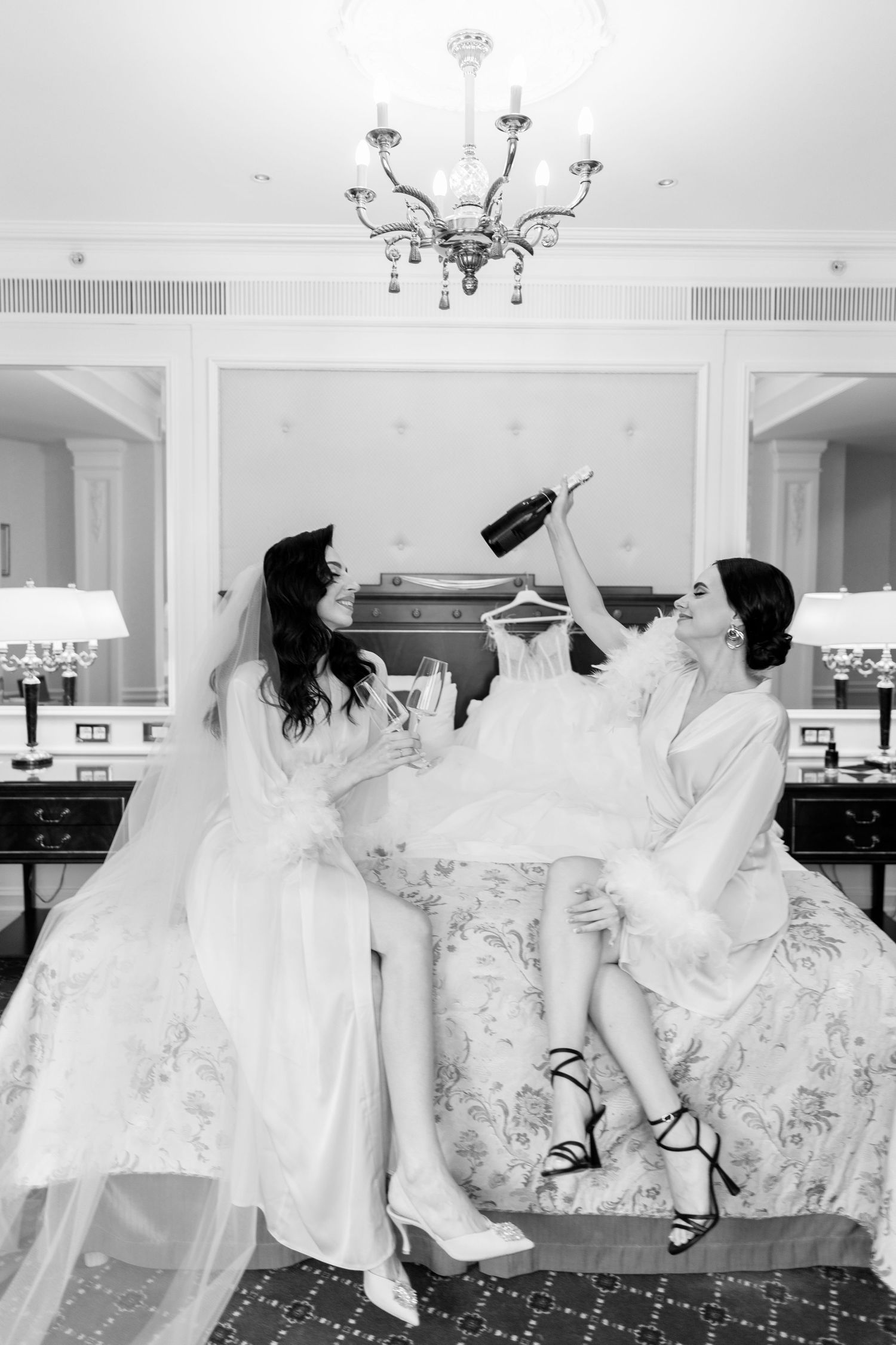 Two brides in white robes celebrate with champagne on a bed in an elegant hotel room, black and white photo.
