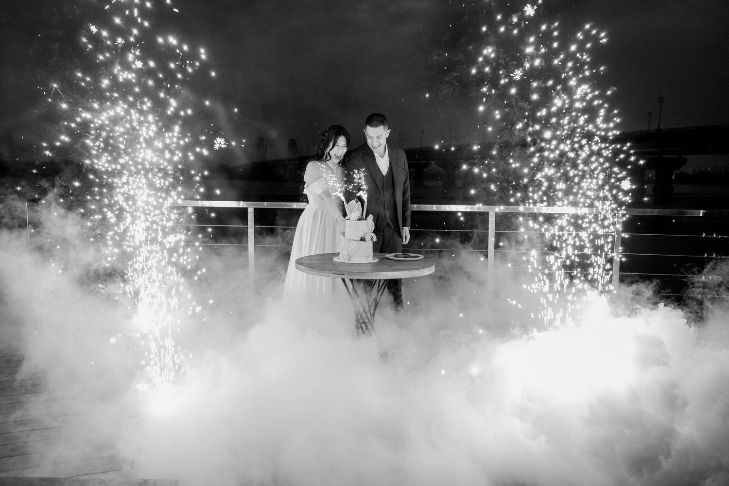 Black and white wedding photo of couple cutting cake surrounded by sparkling fireworks and dramatic fog at night.