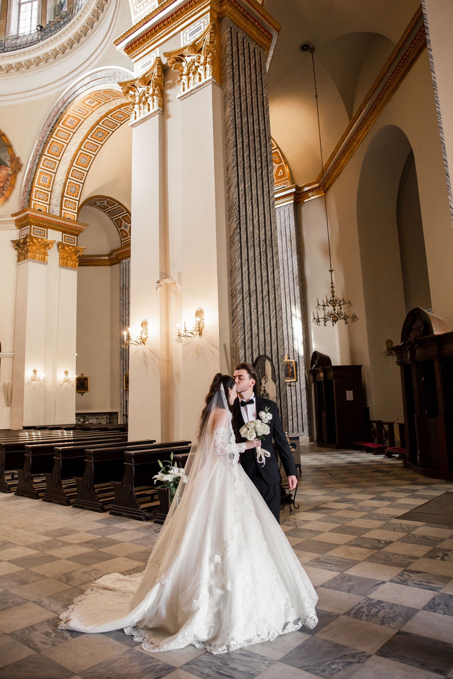 Bride and groom share a romantic kiss inside an elegant church with marble columns and gold ornate architecture.