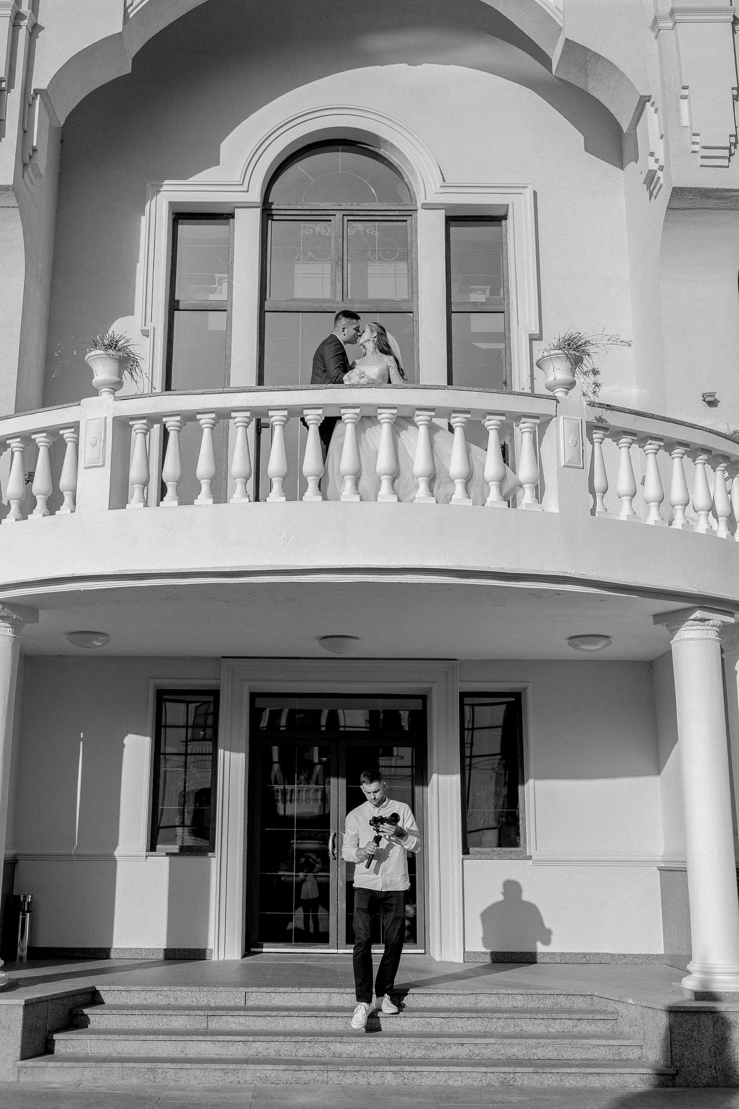 Black and white wedding photo of couple on balcony with man holding bouquet on steps below elegant building.