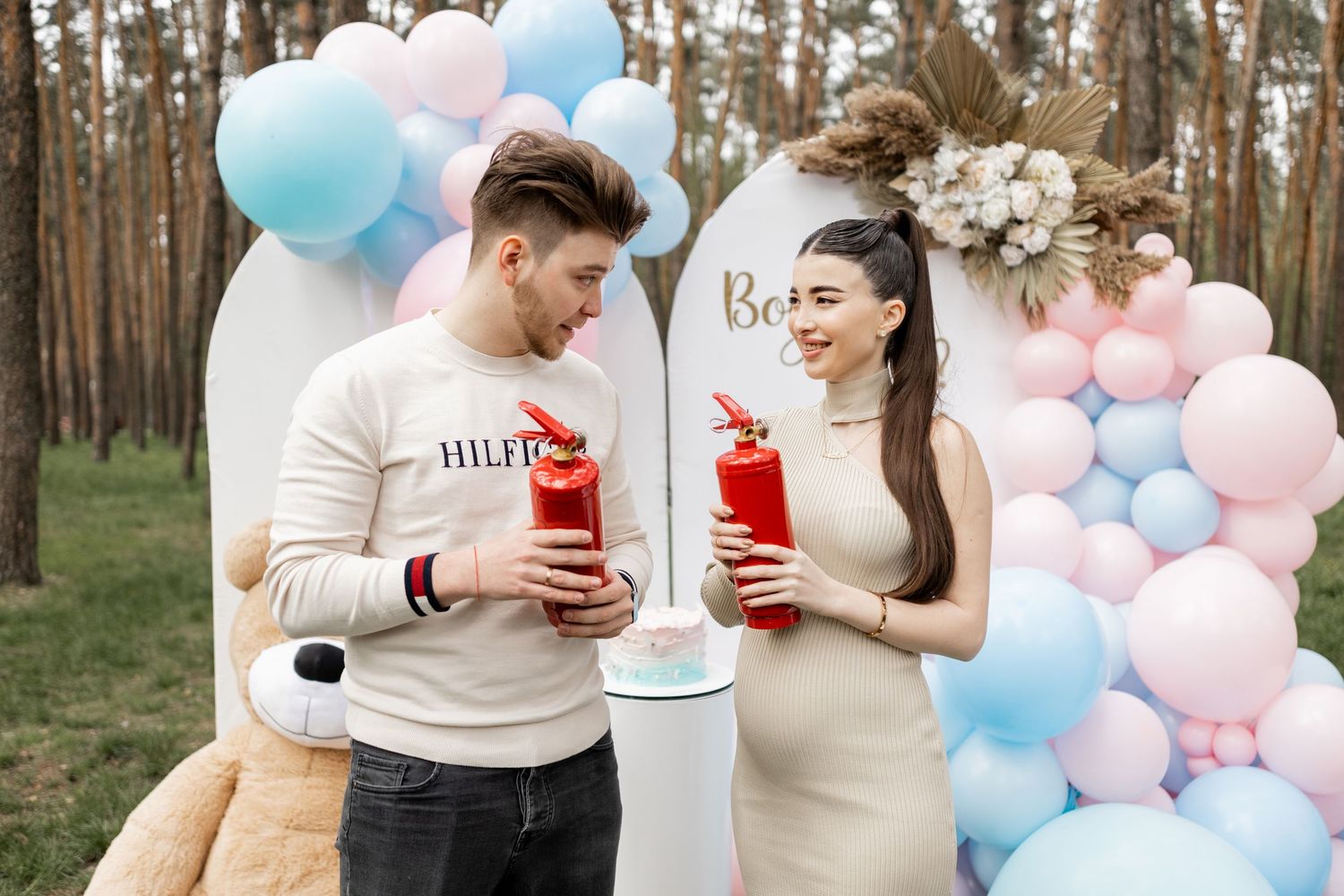 Couple celebrating at gender reveal party with drinks and balloon decorations in forest setting.