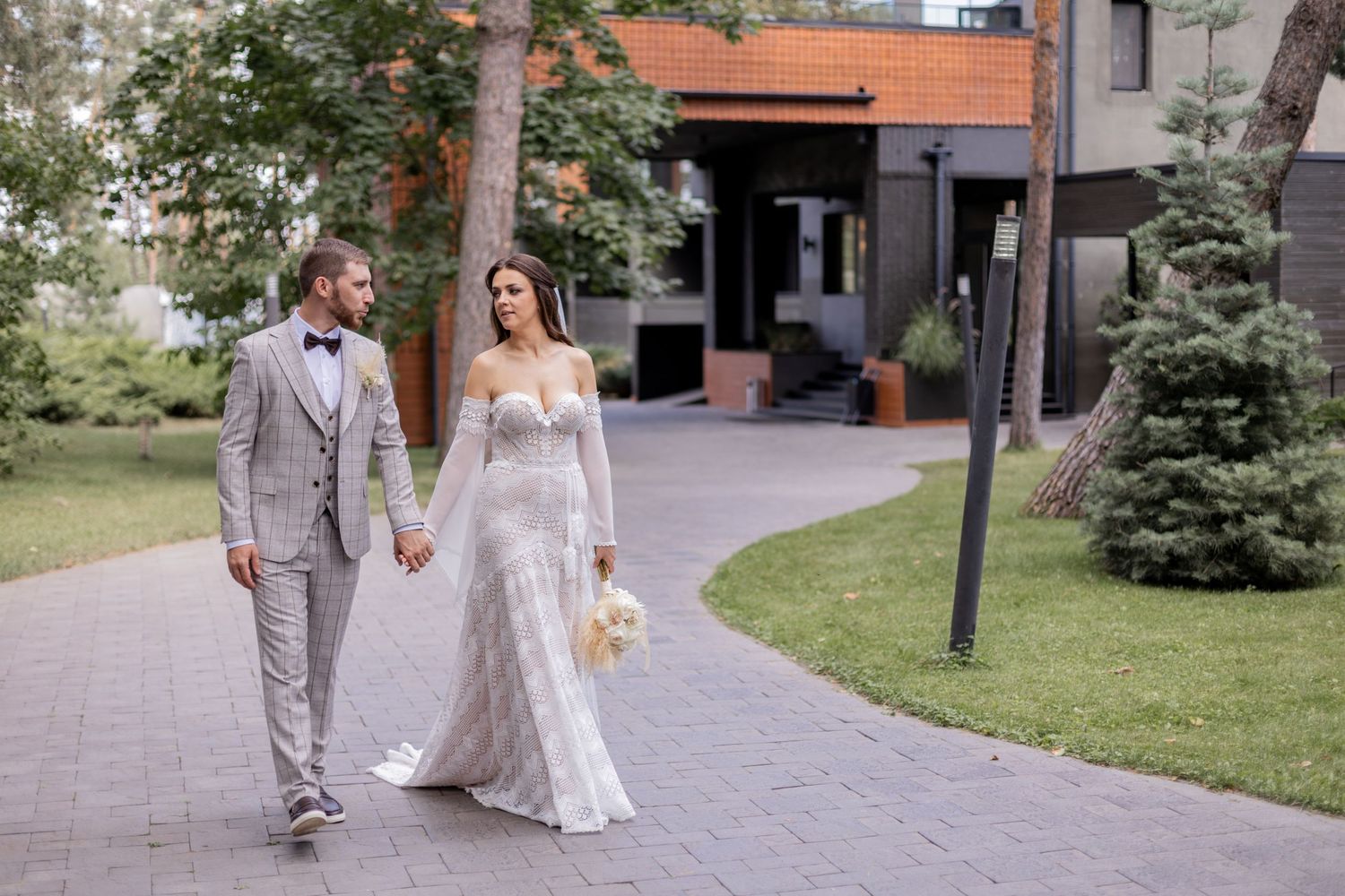Wedding couple strolls along urban path surrounded by evergreen trees.