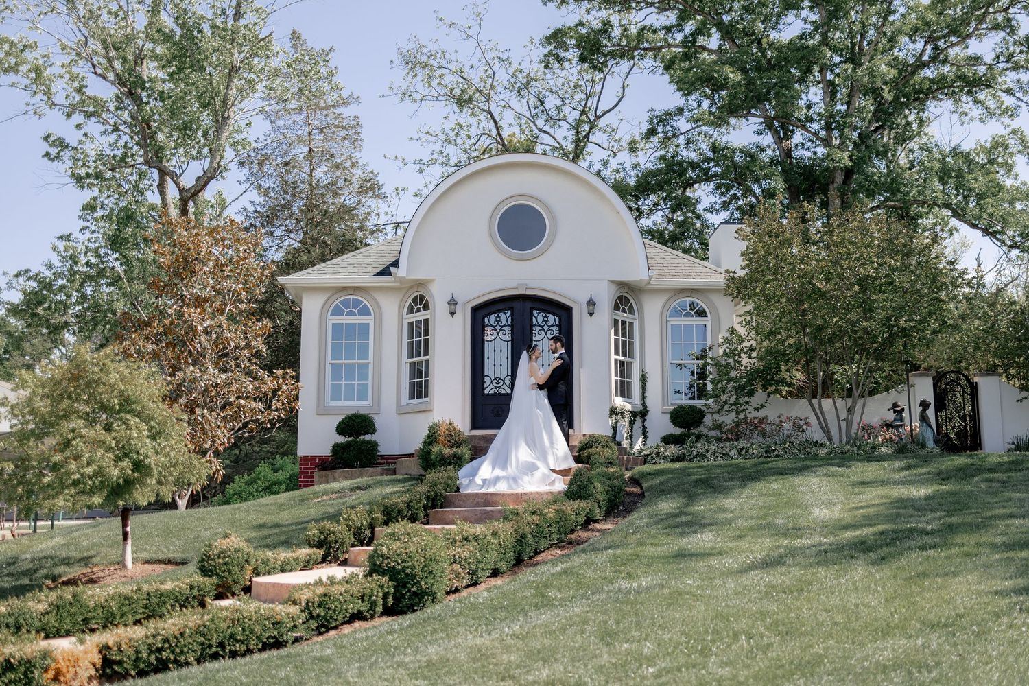 Bride in white wedding gown posing in front of elegant white chapel with arched windows and lush green landscaping.