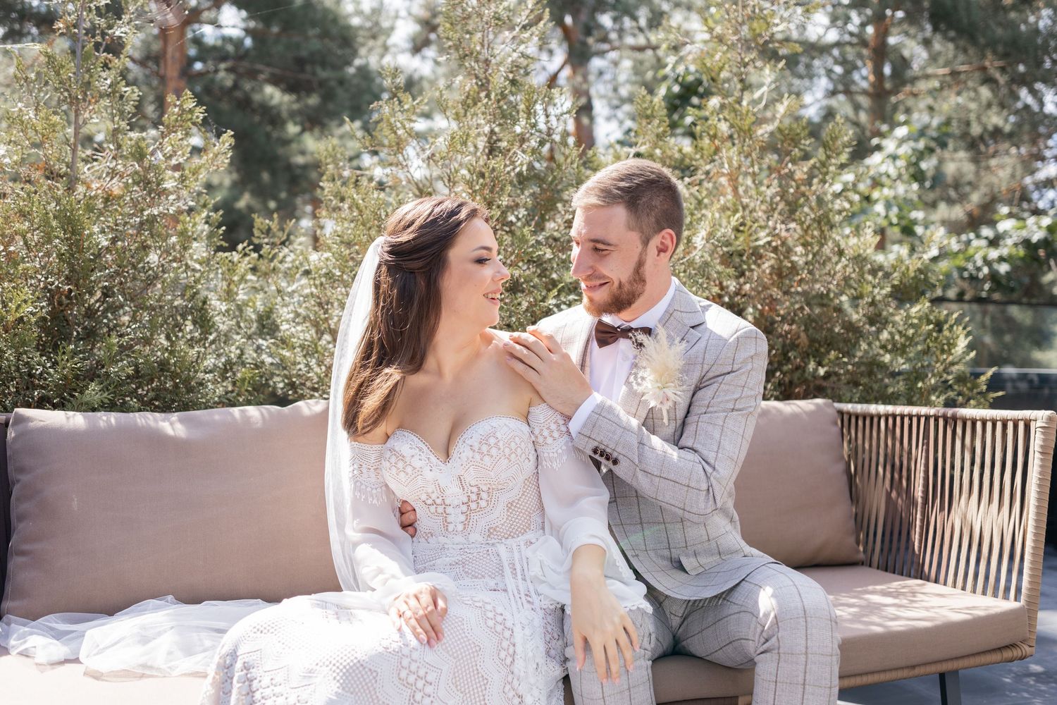 A romantic outdoor photoshoot featuring a couple in wedding attire sitting on a bench surrounded by trees.
