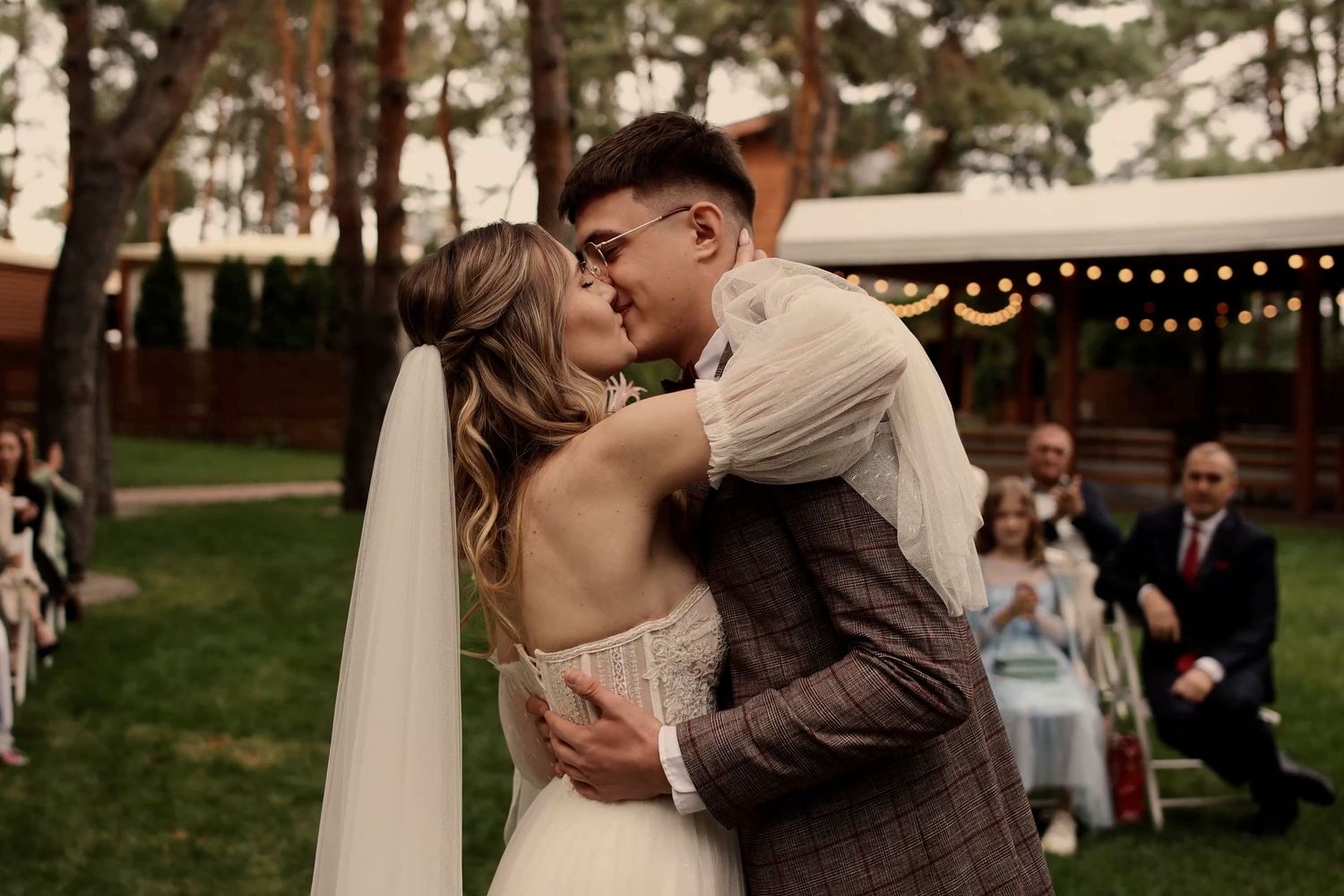 Bride and groom share first kiss at outdoor wedding ceremony with guests and string lights in background.