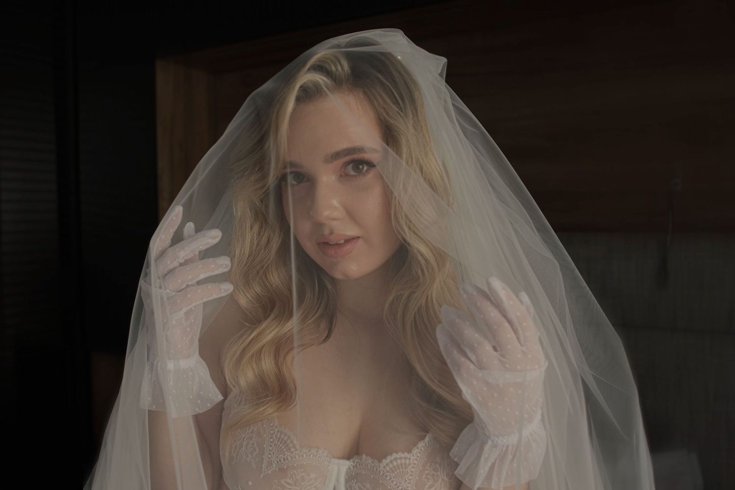Blonde bride wearing a white veil and lace corset with white gloves in a softly lit room.