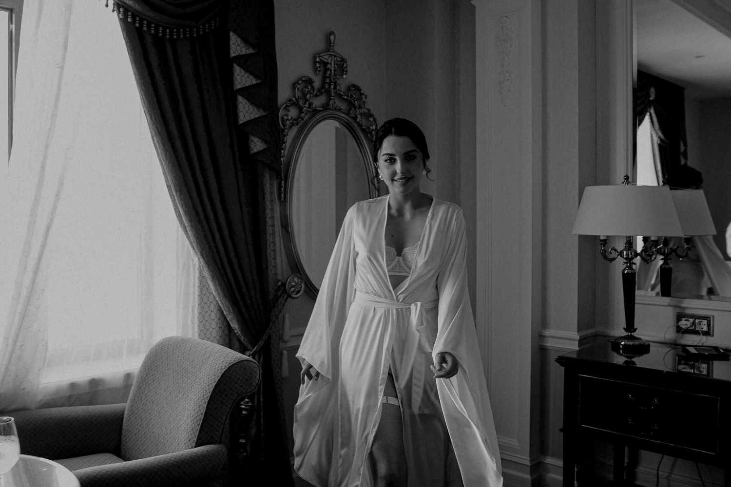 Bride in white flowing robe standing by window in elegant room, black and white wedding preparation photo.