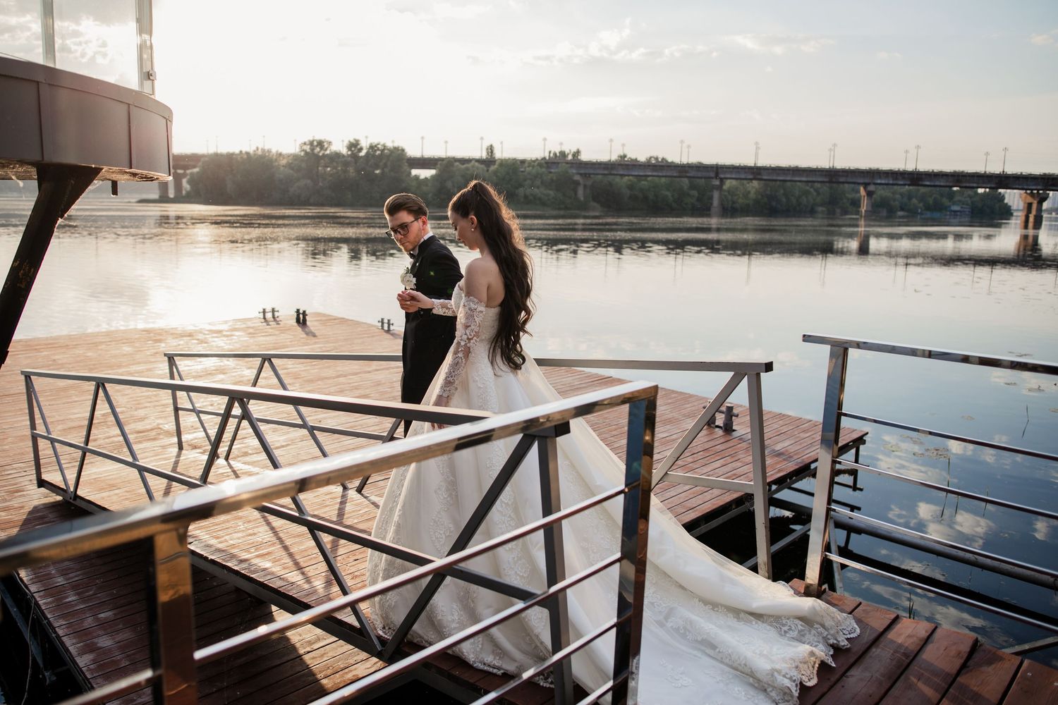 Bride and groom share a romantic moment on a wooden dock by a river at sunset.