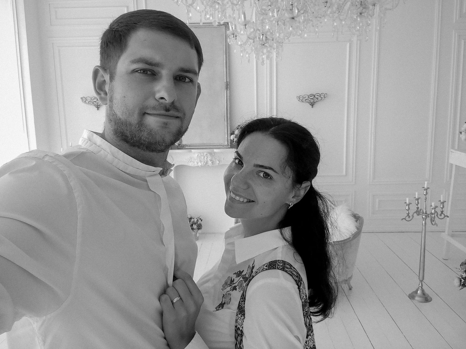 Black and white selfie of a smiling couple in an elegant white room with chandelier.