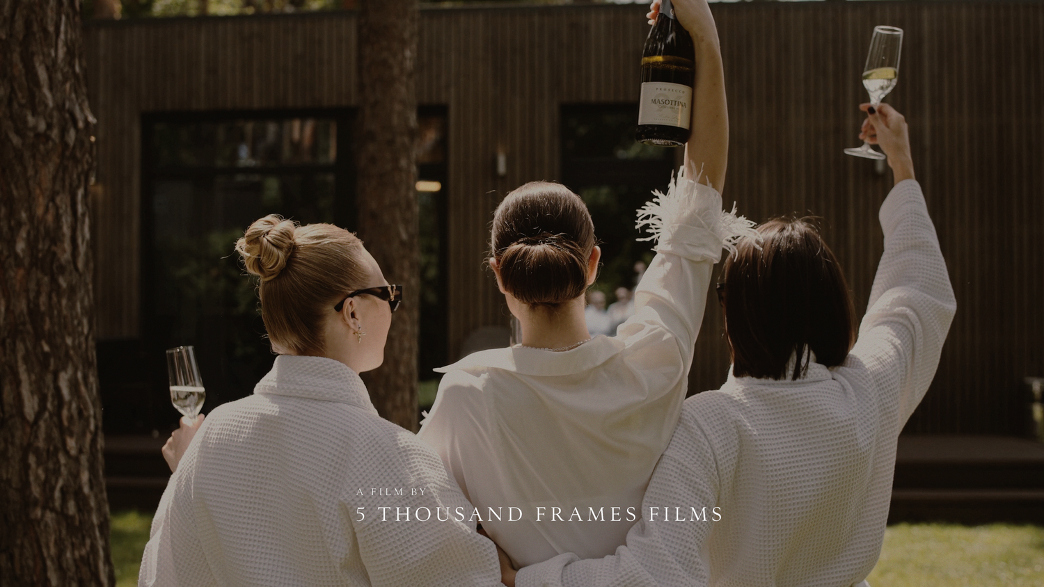 Three women in white robes raise champagne glasses outdoors, celebrating a wedding day with 5 Thousand Frames Films.