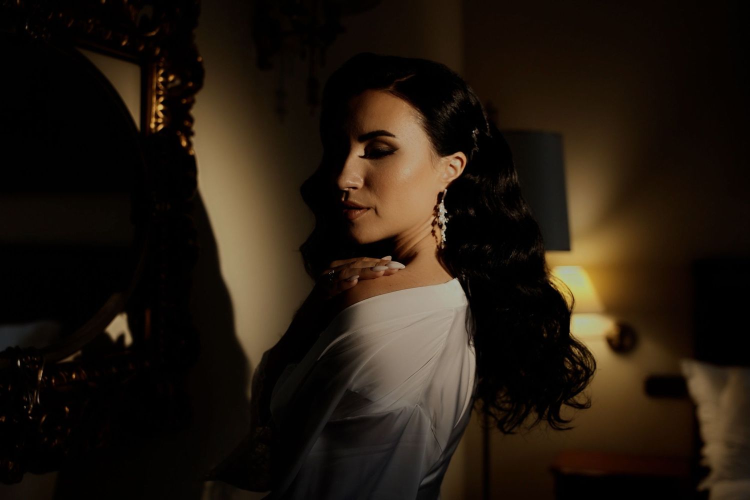 Elegant woman in white off-shoulder dress with ornate earrings, posed in moody dramatic lighting.