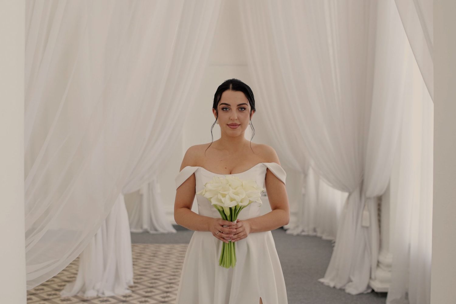 Bride in off-shoulder white gown holding calla lily bouquet stands in elegant white draped venue.