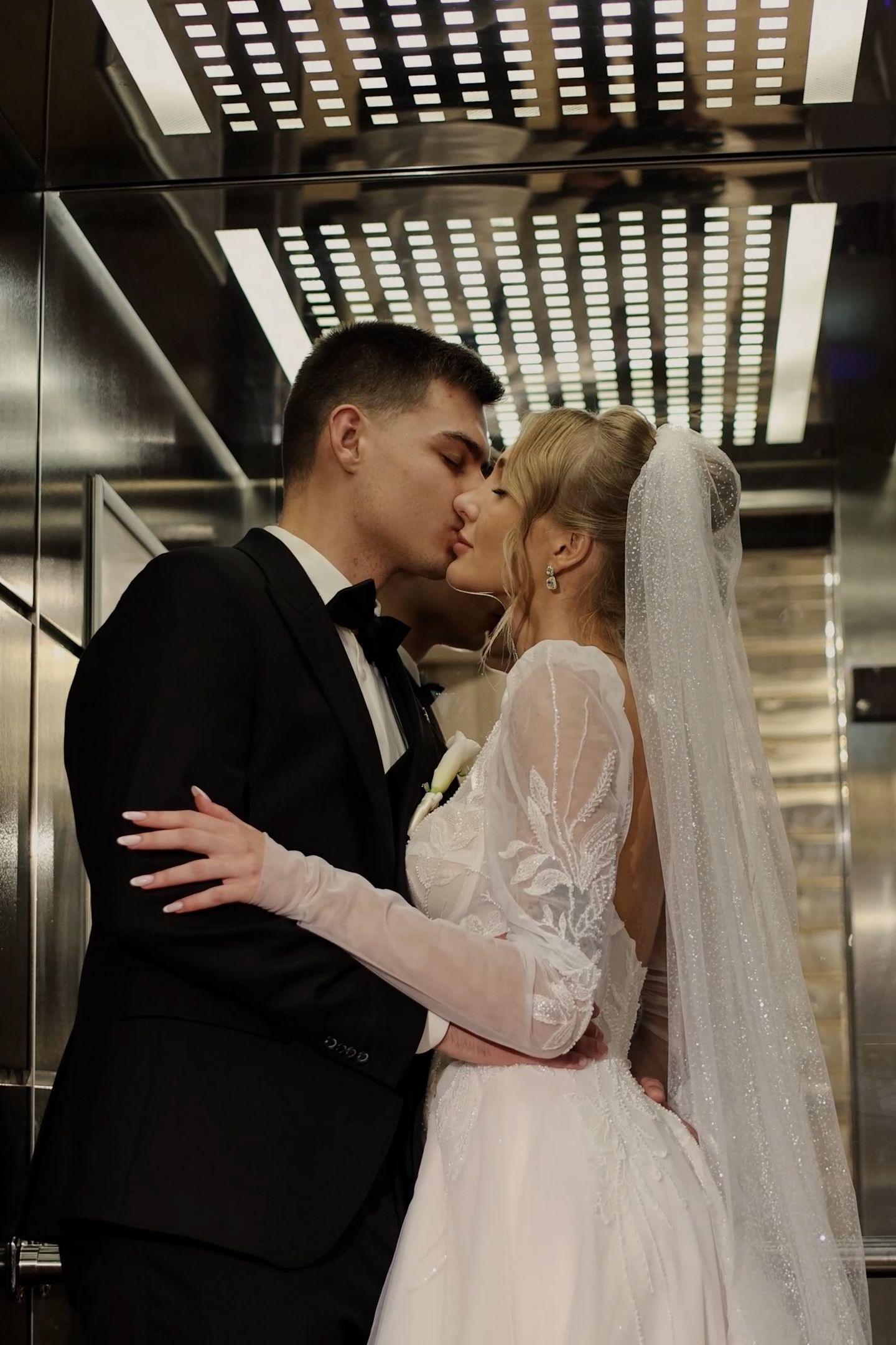 Newlywed couple sharing a romantic kiss in an elevator, bride in white gown with veil, groom in black tuxedo.