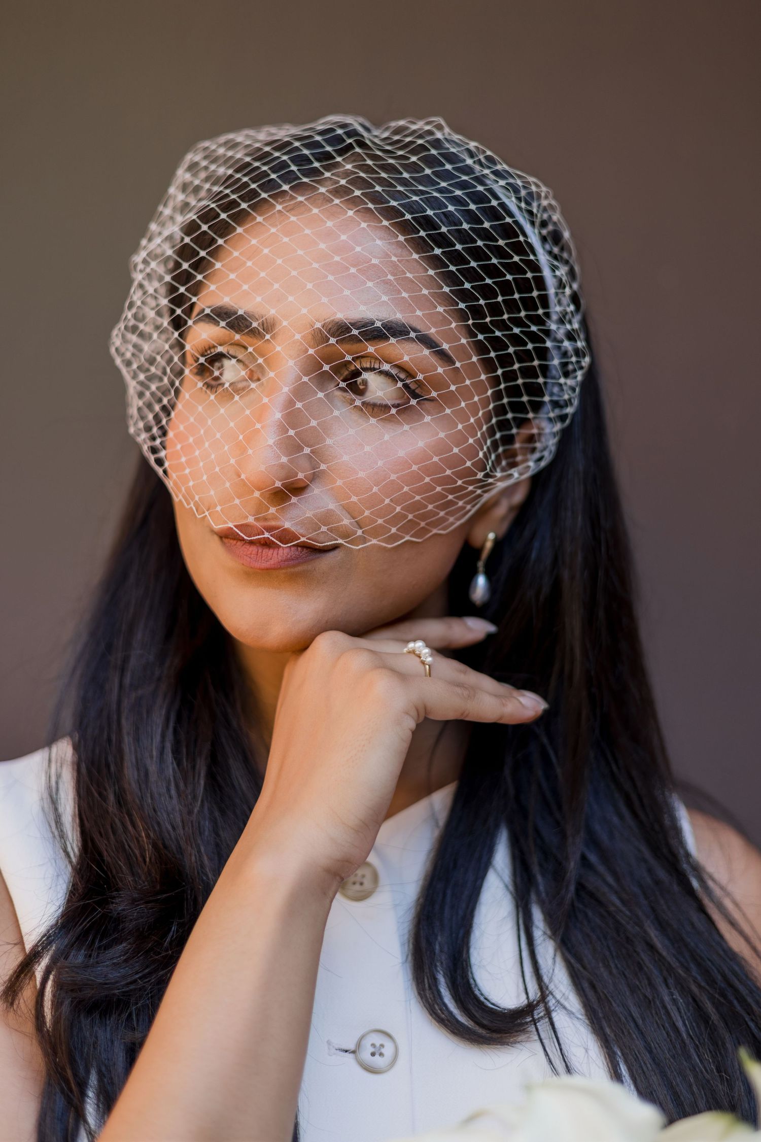 Bride wearing a delicate white birdcage veil and white dress, posing thoughtfully with hand on chin.