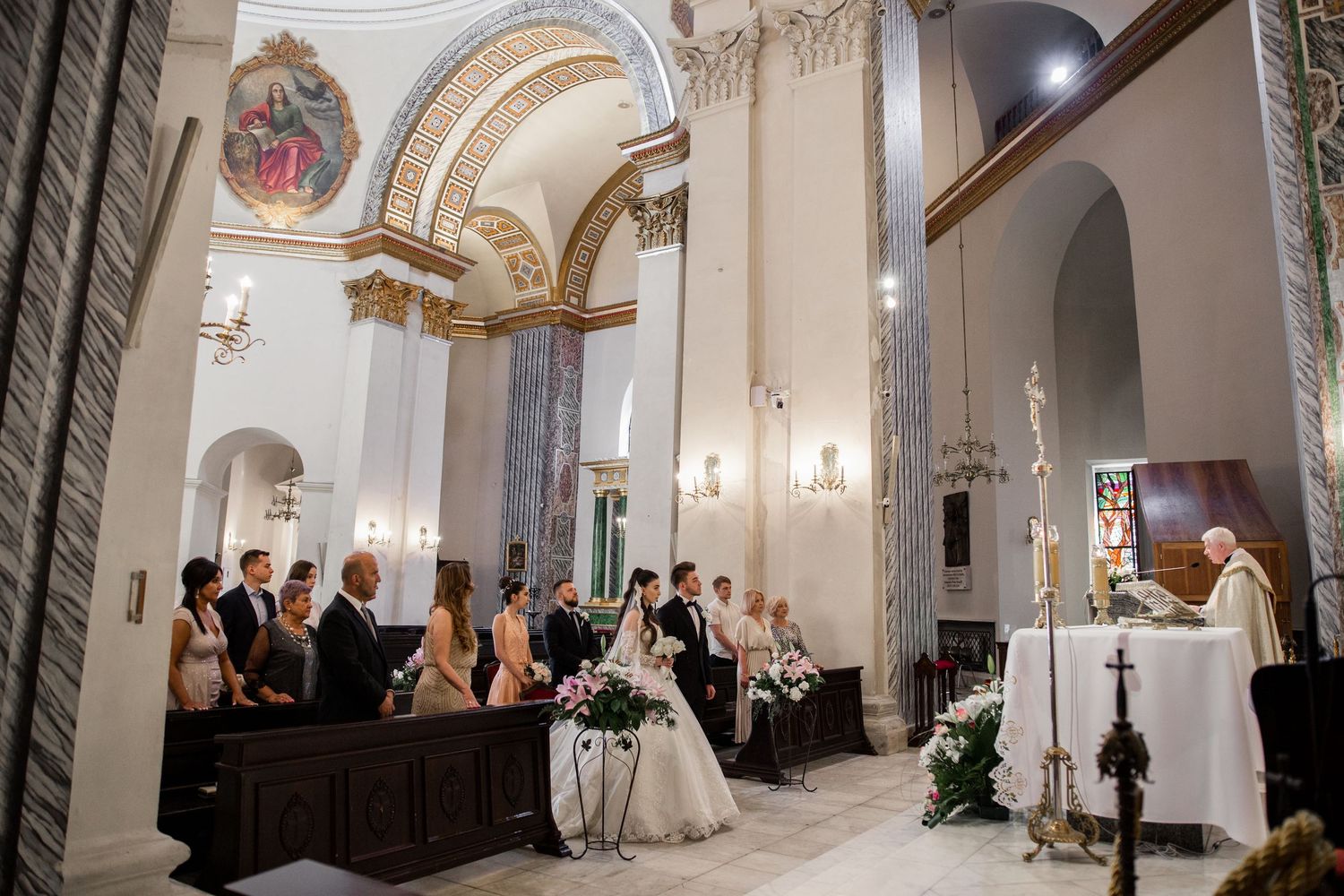 A wedding ceremony in an elegant white church with the bride, groom, wedding party, and priest gathered at the altar.