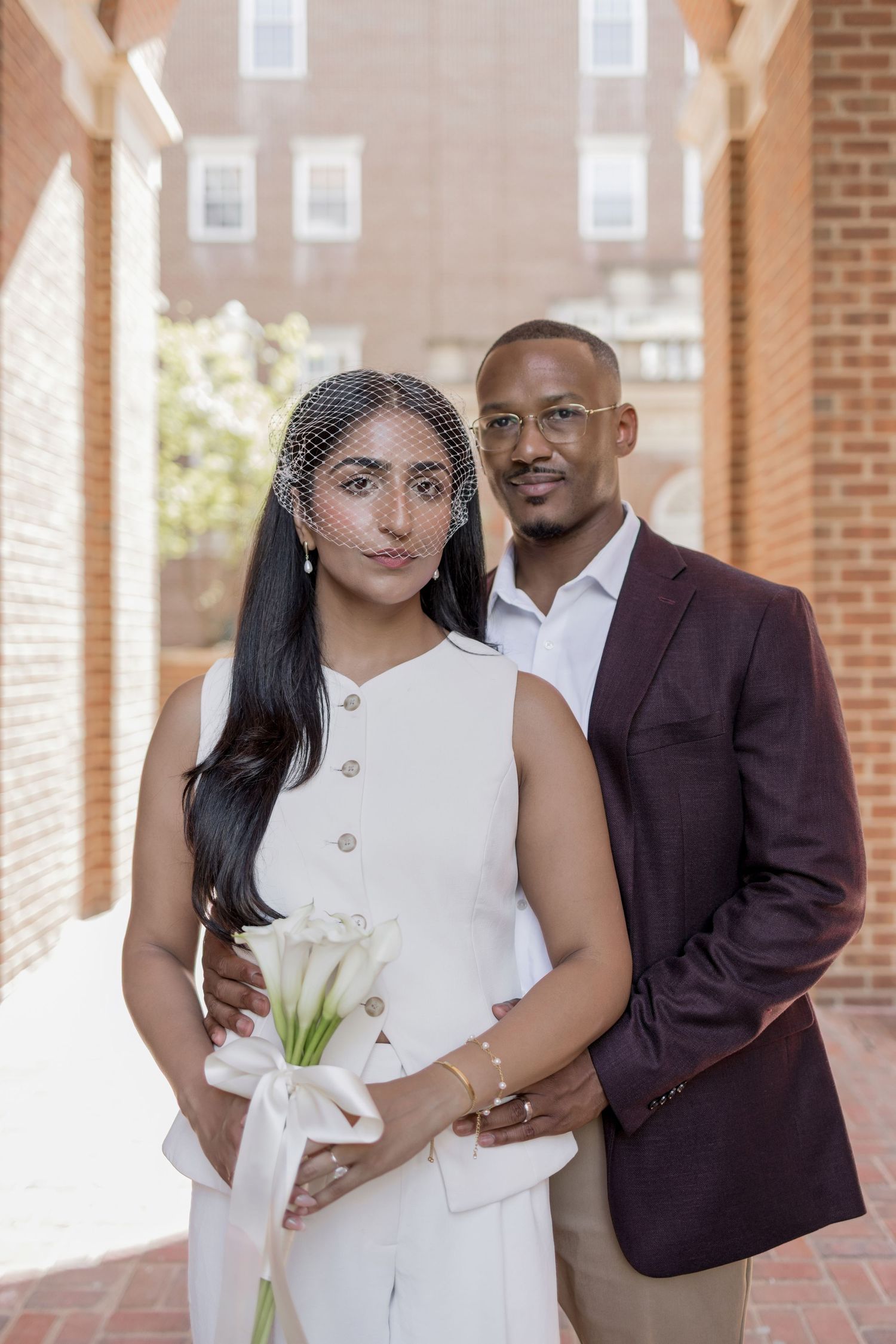 Elegant couple posing for wedding portrait, bride in white jumpsuit with veil holding calla lilies, groom in burgundy blazer.