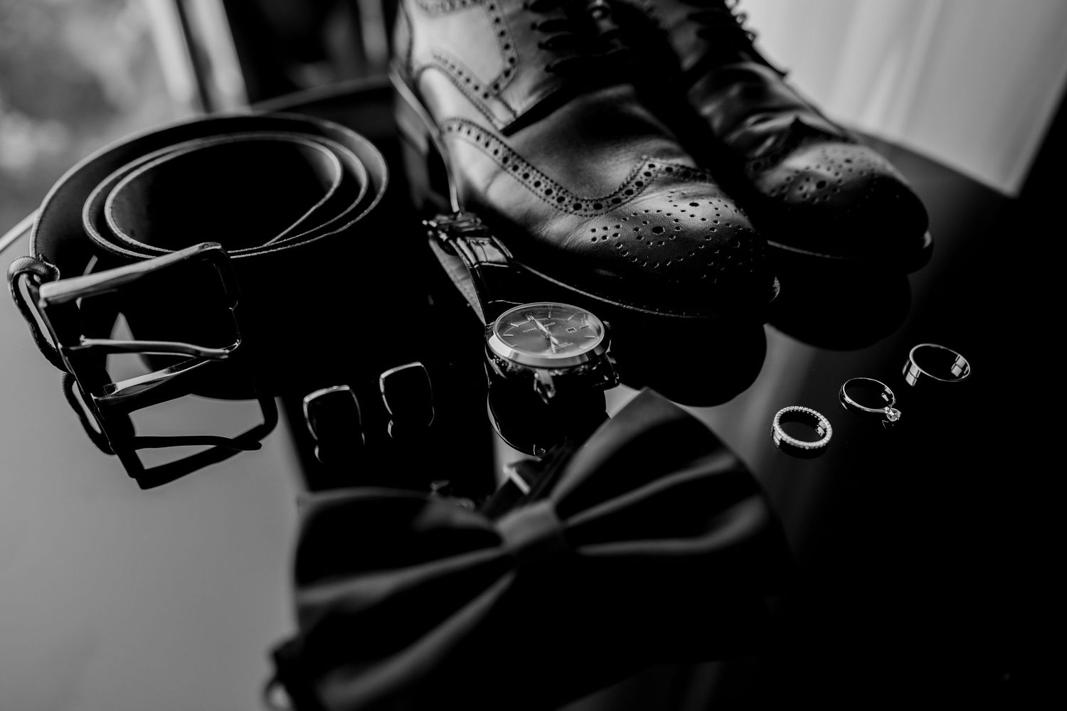 Black and white flat lay of mens wedding accessories including dress shoes, belt, watch, bow tie, and rings.