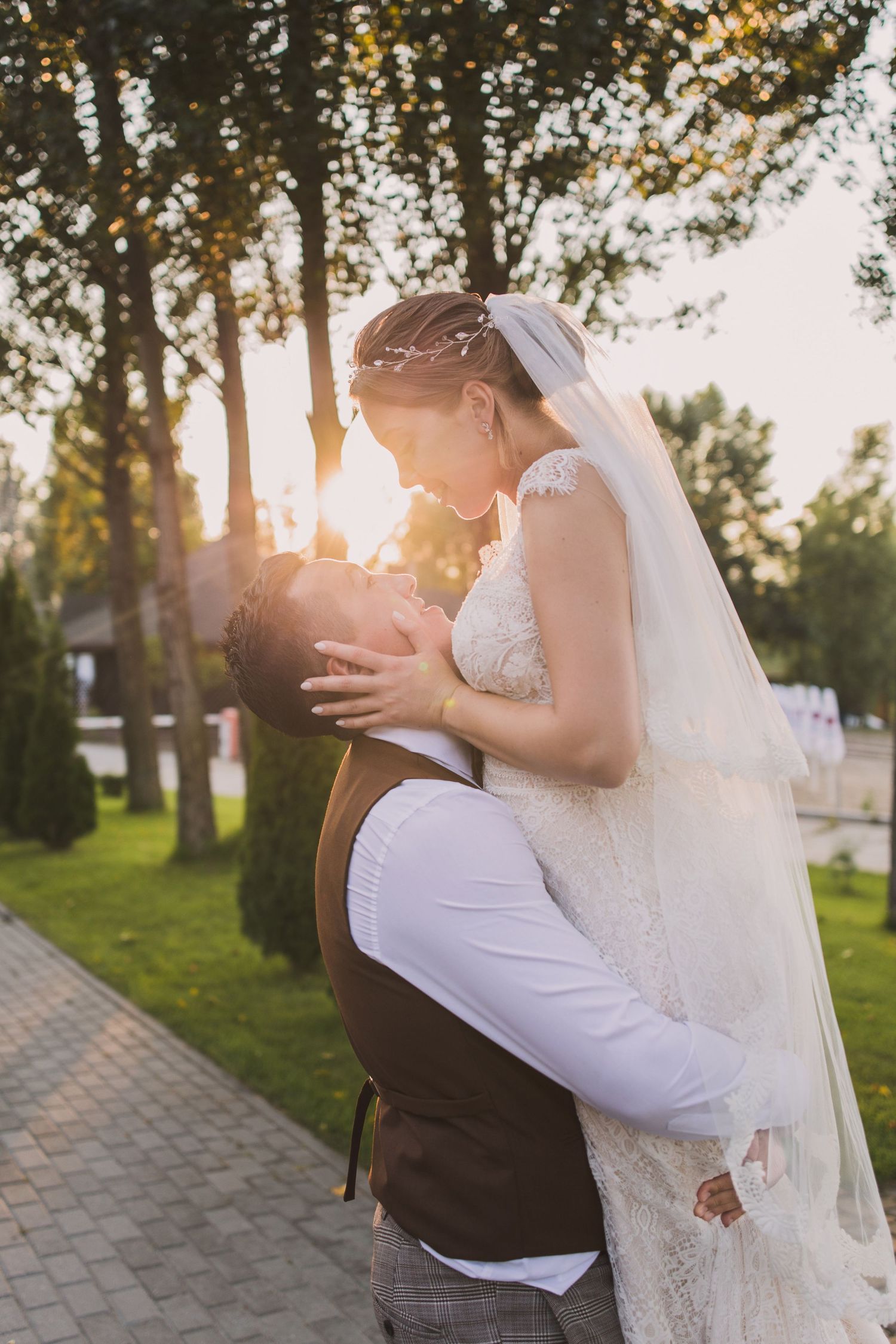Bride and groom sharing a romantic sunset kiss outdoors, surrounded by lush green trees in a beautiful wedding setting.