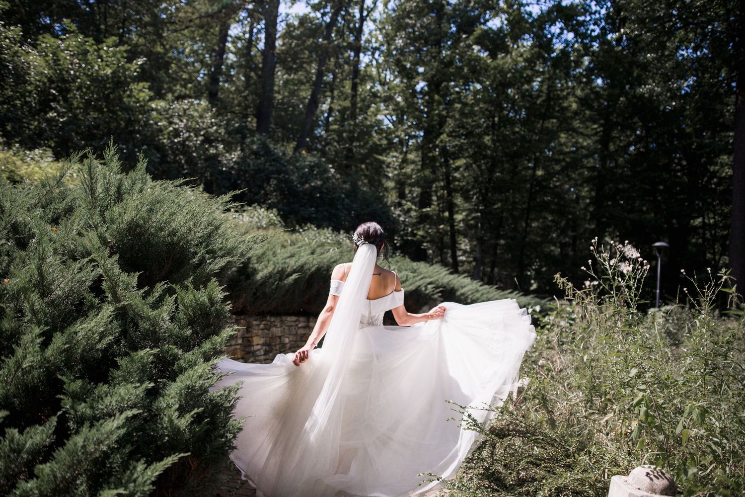 Bride in flowing white wedding gown and veil walks through lush green garden surrounded by tall trees.
