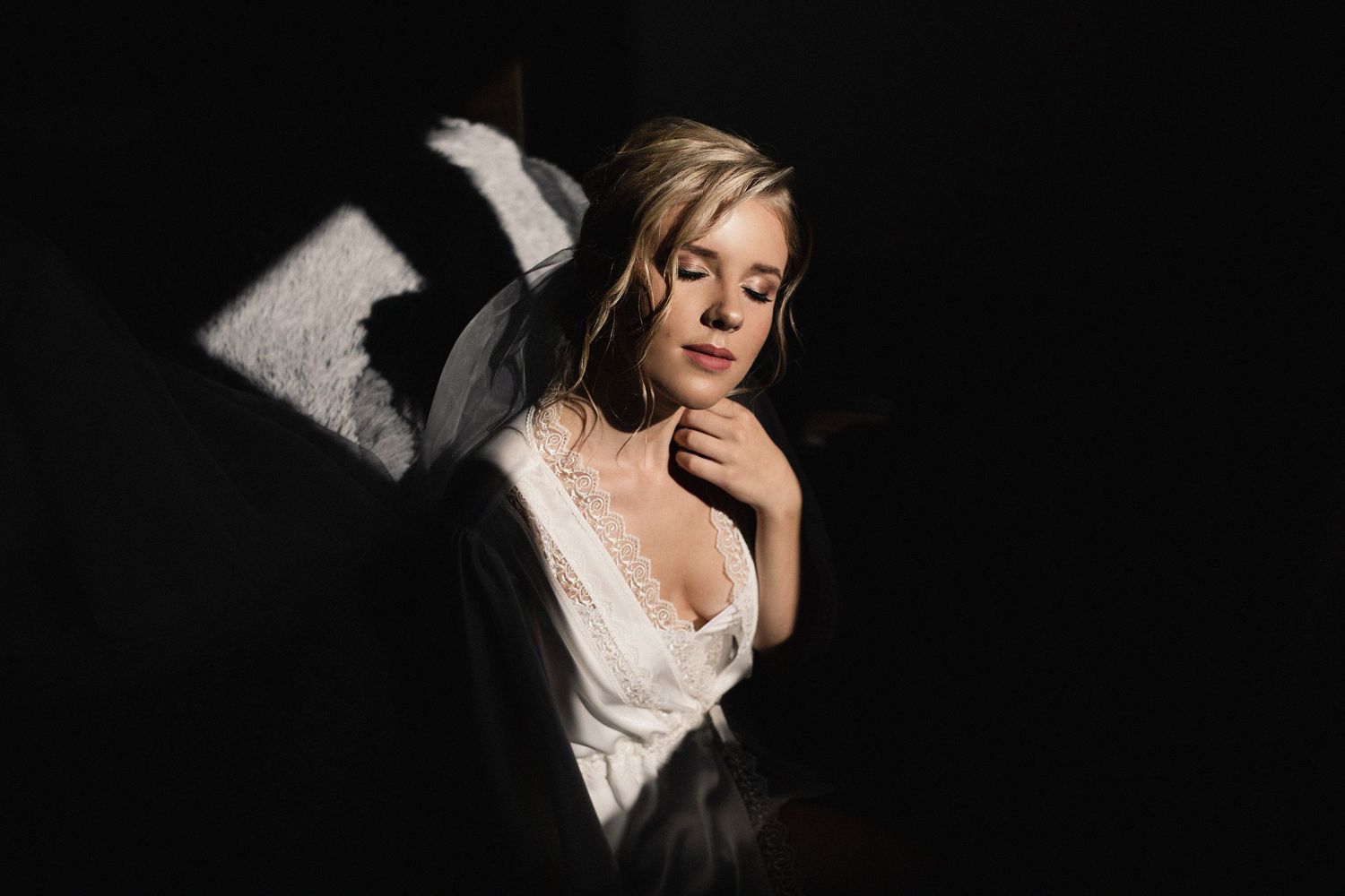 Blonde woman in white lace robe poses in dramatic chiaroscuro lighting against a dark background.