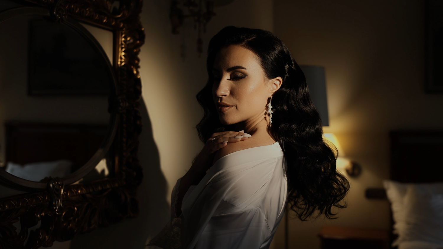 Elegant woman in white off-shoulder top with dangling earrings posing in dimly lit room near ornate gold mirror.