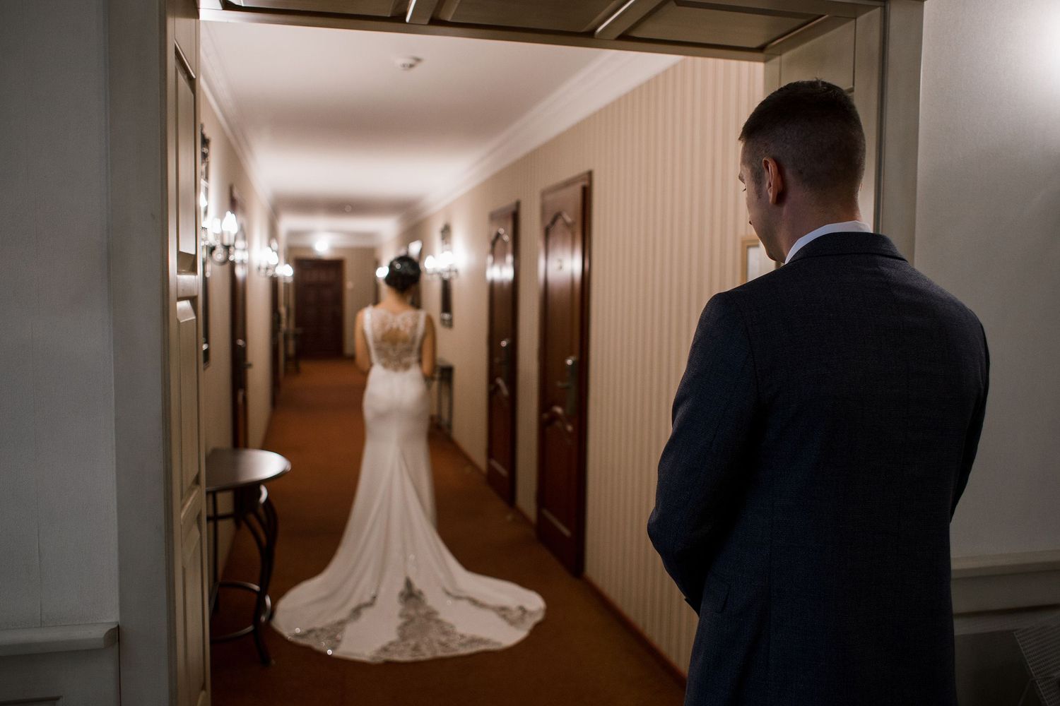 Bride in elegant lace wedding gown with train stands in hotel hallway as groom in navy suit looks on.