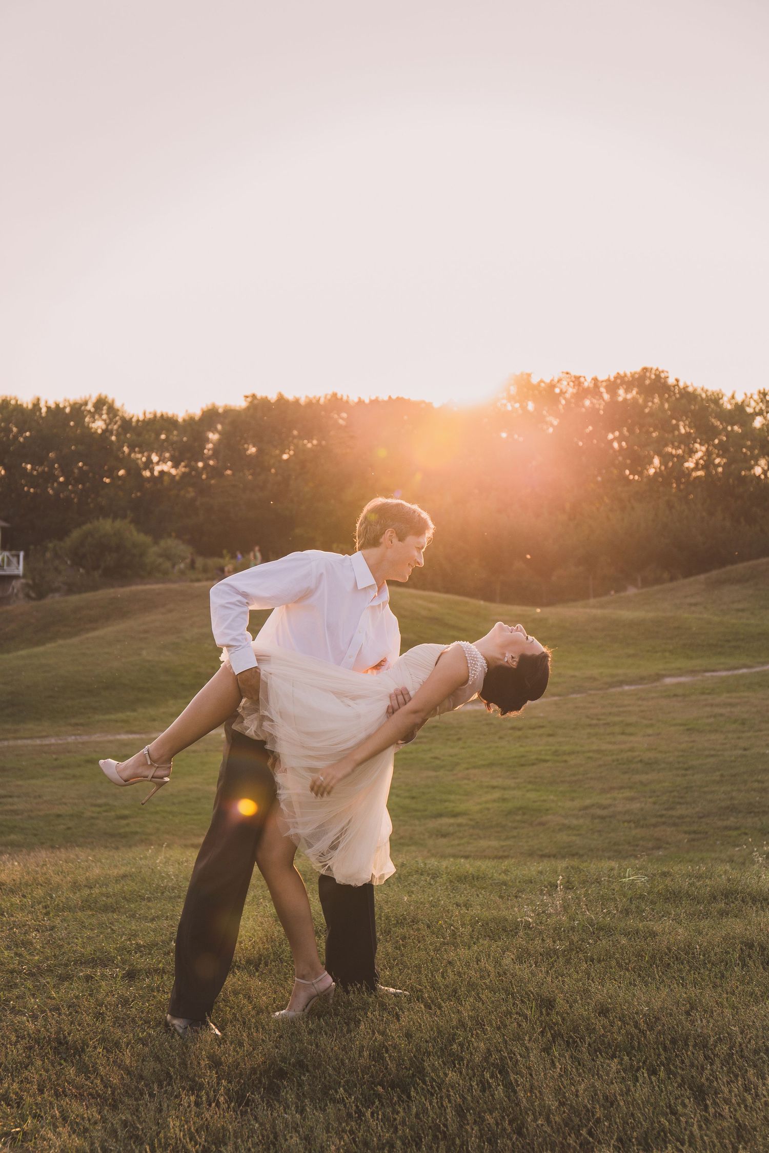 A couple shares a romantic dance at golden hour sunset on a lush green field, with the man lifting his partner gracefully.