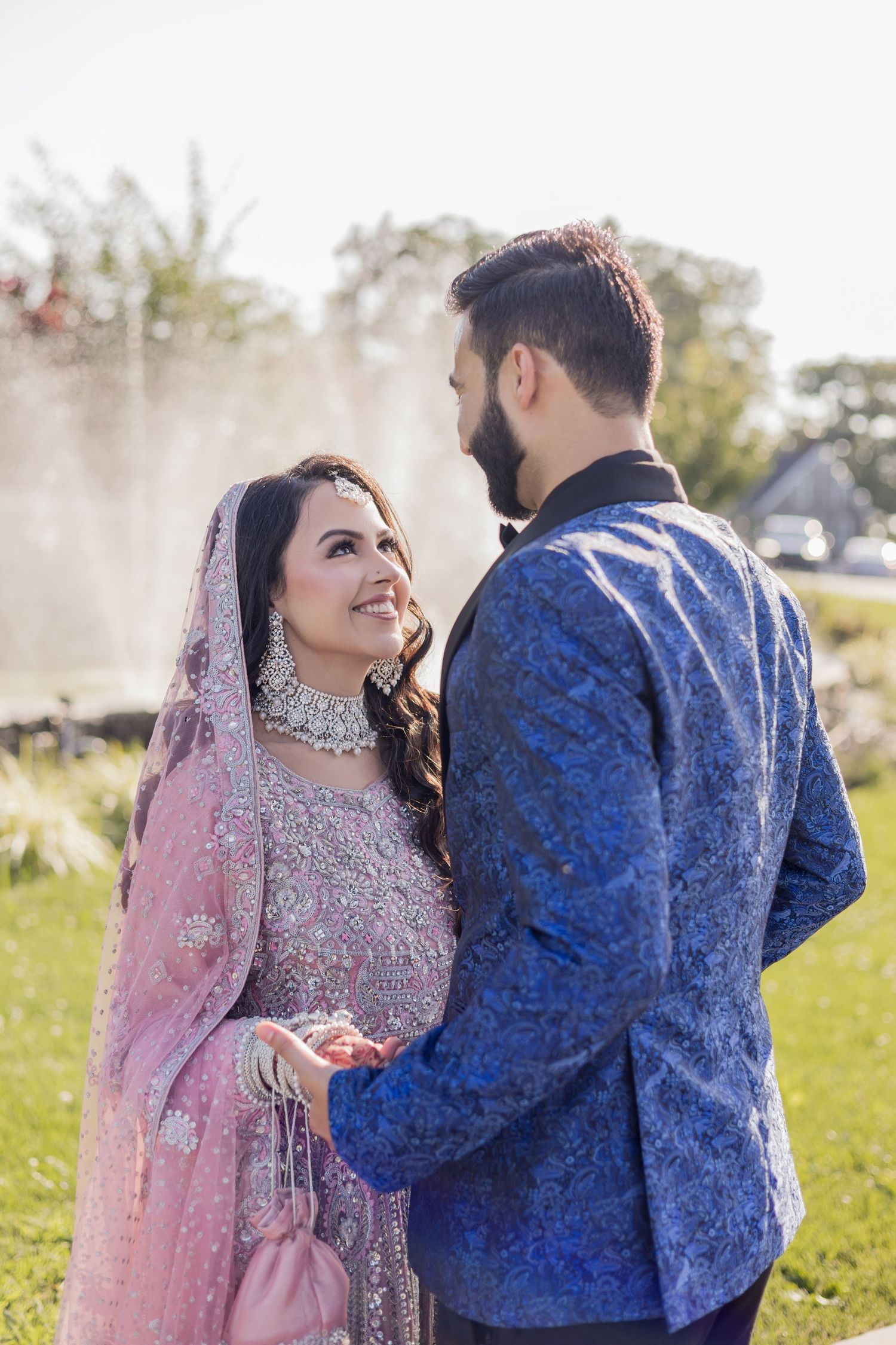 South Asian couple in traditional wedding attire, bride in pink lehenga and groom in blue sherwani, holding hands outdoors.