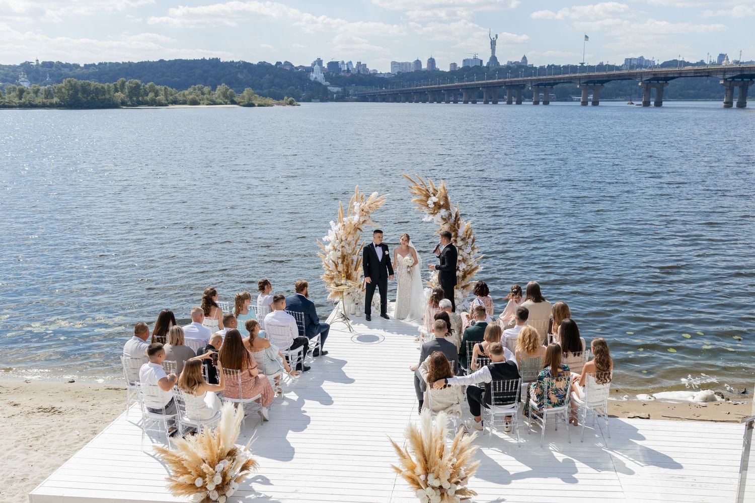 Outdoor waterfront wedding ceremony on white dock with floral arch, guests seated, bridge and city skyline in background.