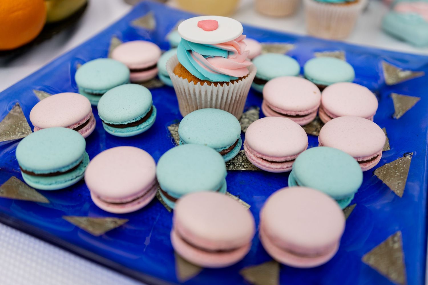 Pink and blue macarons arranged on a blue serving plate with a decorated cupcake.