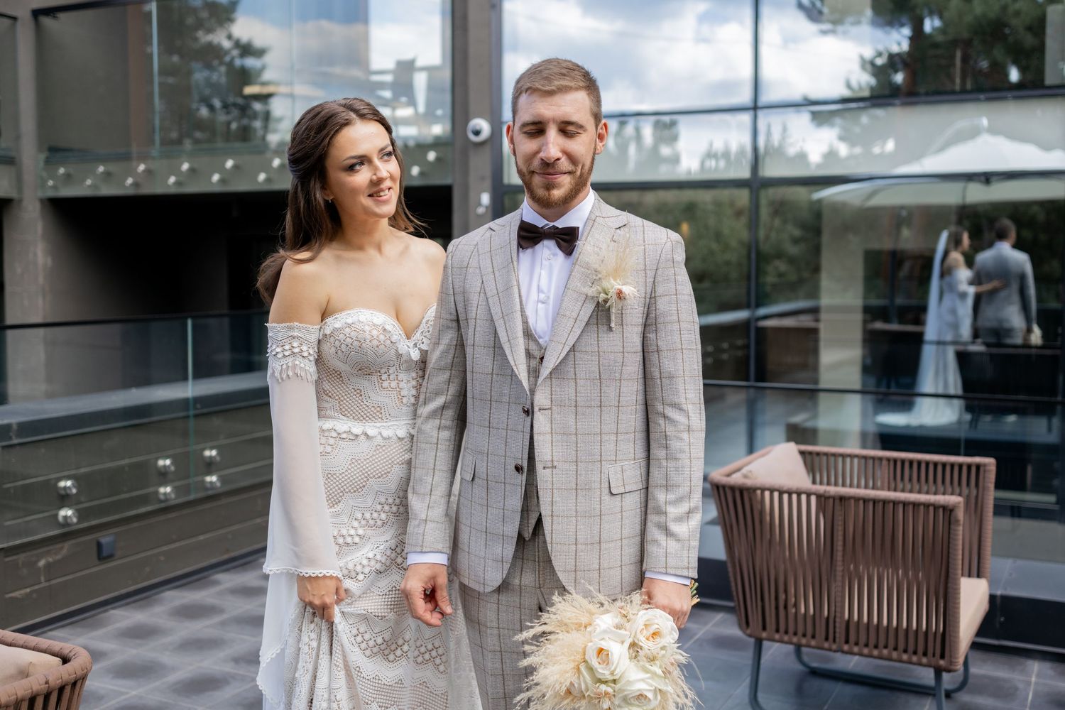 A couple in elegant wedding attire share a romantic moment on a modern balcony with glass railings.