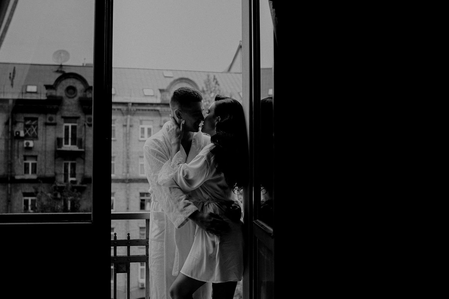 Black and white photo of a romantic couple embracing on a balcony with city buildings in the background.