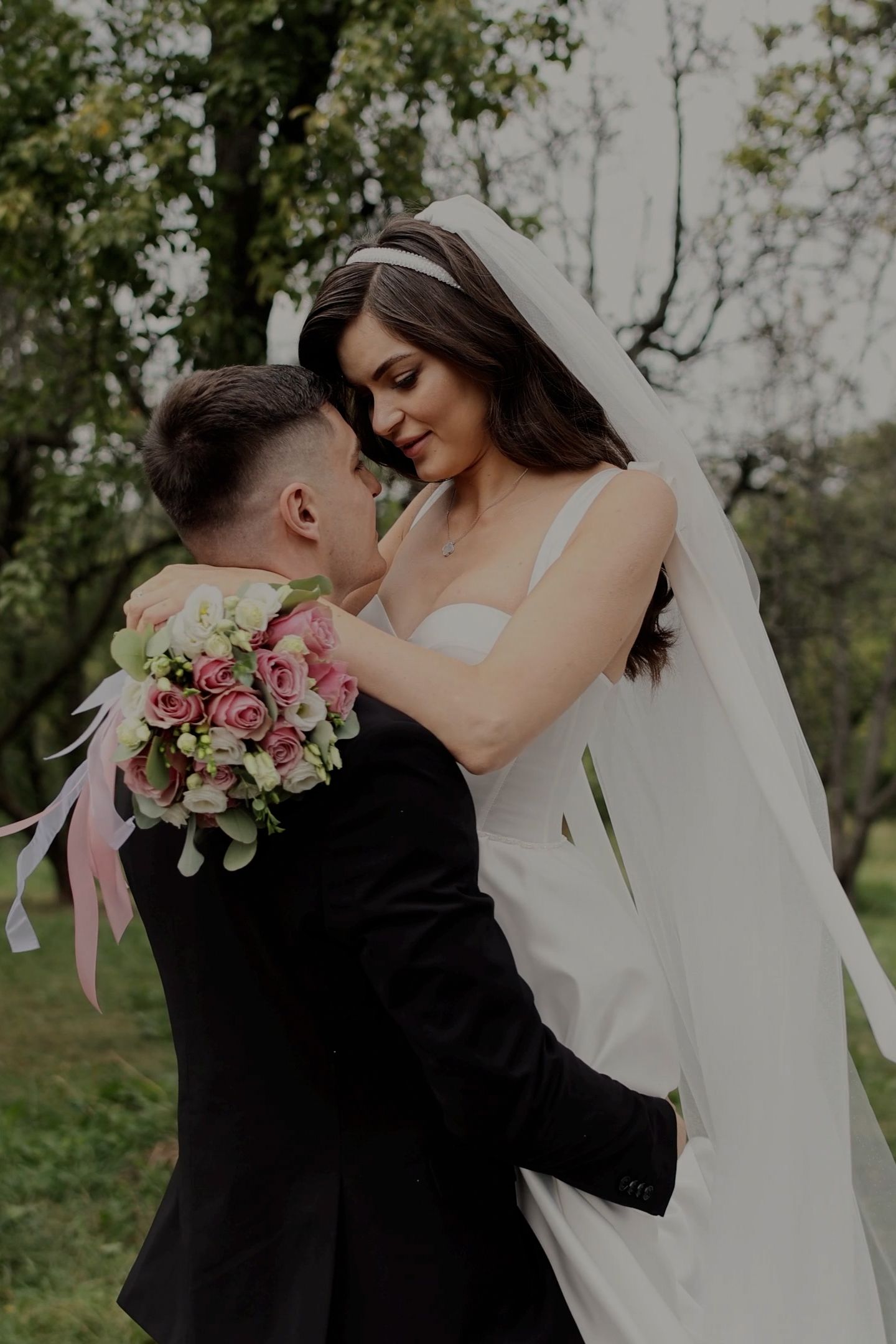 A bride and groom share a romantic moment outdoors, with the bride holding a pink rose bouquet while wearing a white gown and veil.