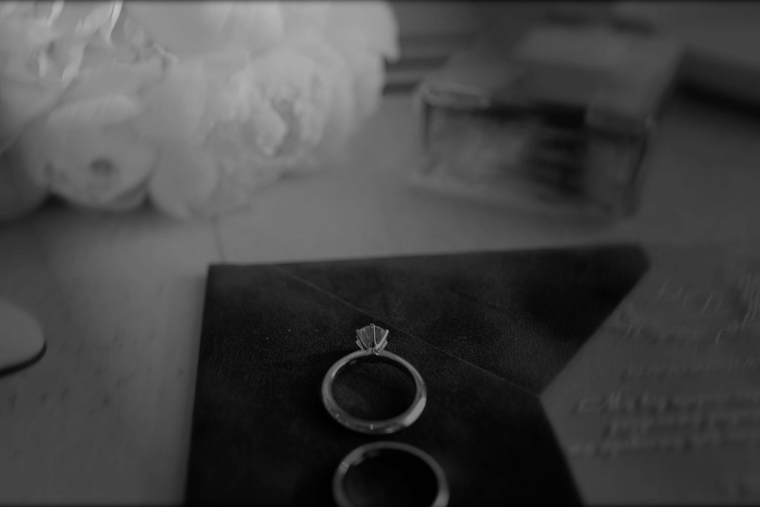 Black and white photo of diamond engagement ring and wedding bands on dark surface with white floral bouquet.
