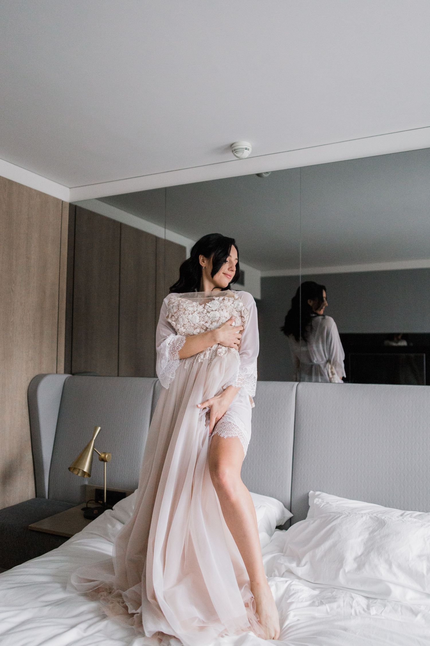 A bride in a sheer lace bridal robe stands on a white bed, with her reflection visible in a large mirror behind her.