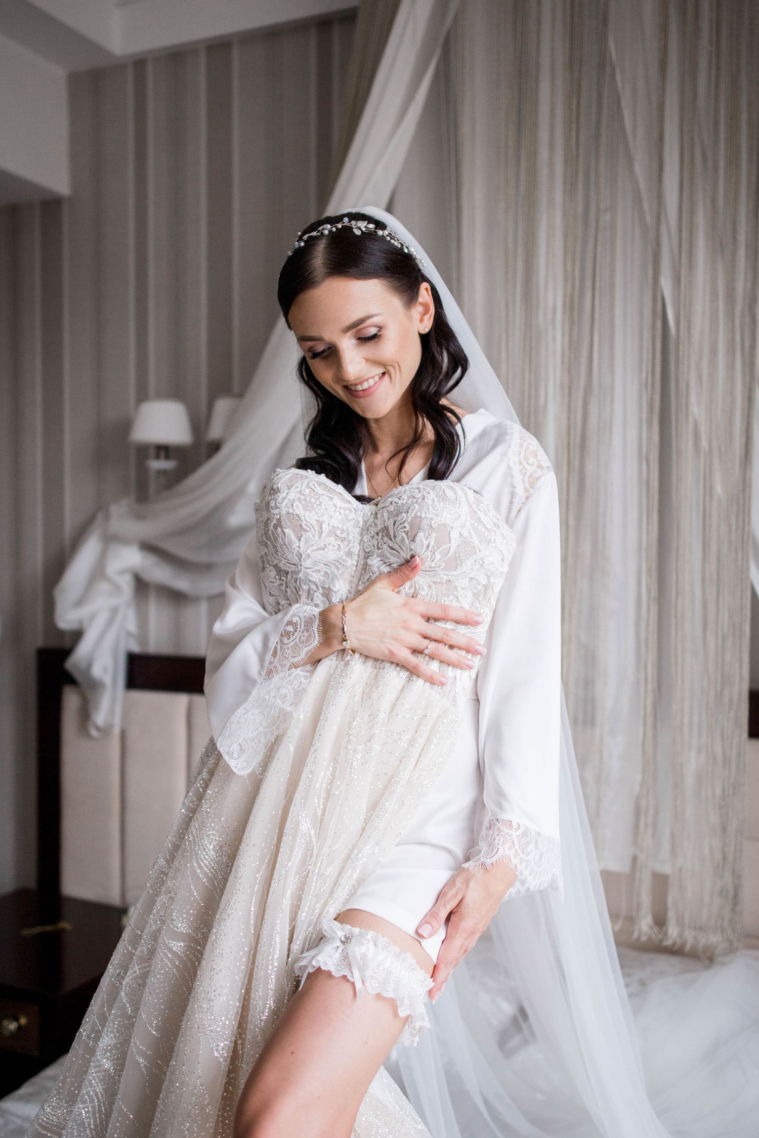 Smiling bride in lace wedding dress and veil adjusting garter in elegant bridal suite.