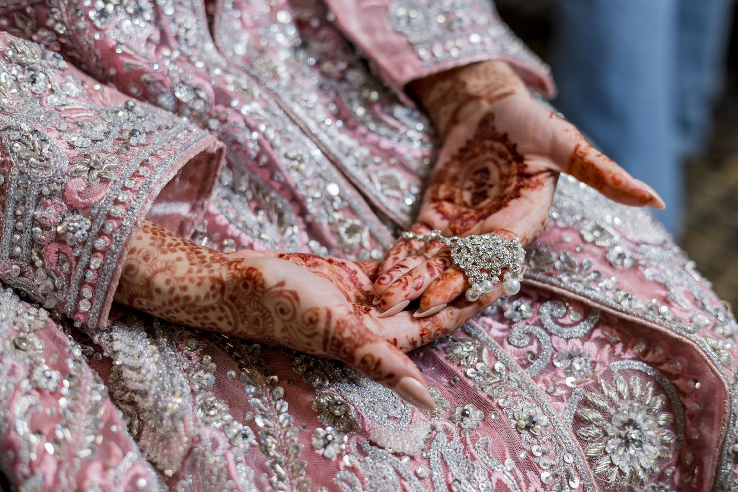 Bride with intricate henna mehndi designs on hands holding ornate jewelry, wearing embroidered pink bridal lehenga.