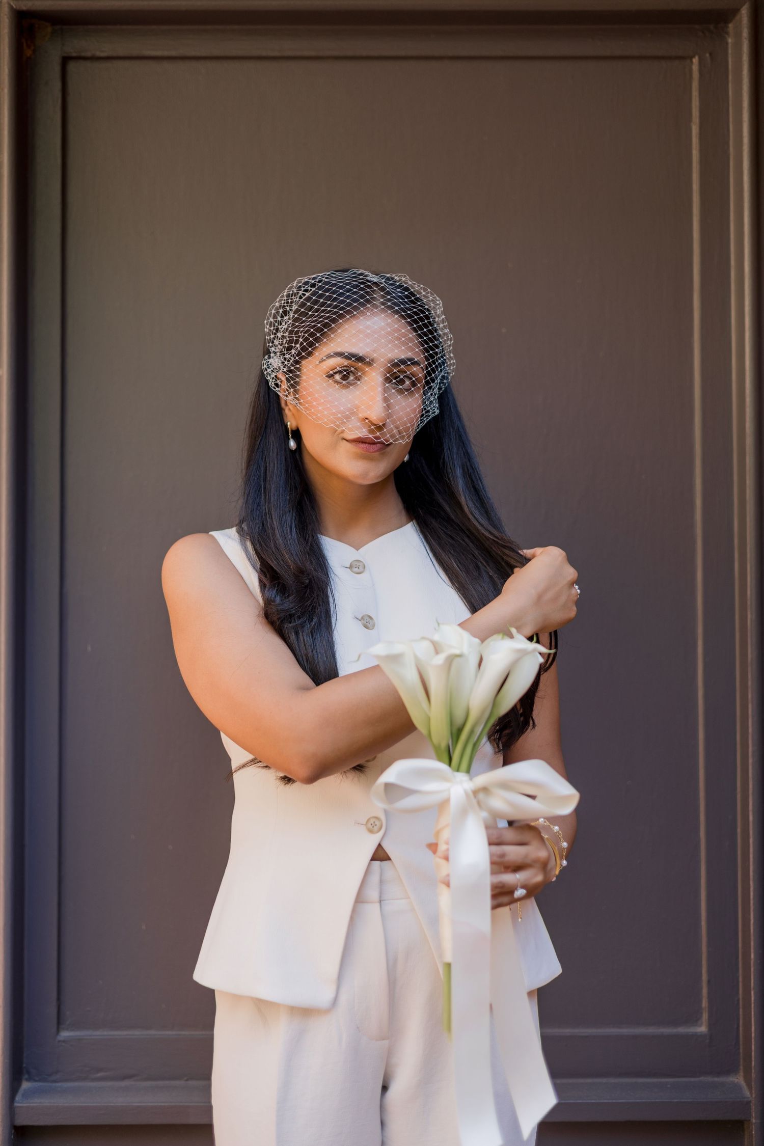 Bride in white pantsuit holding calla lily bouquet with birdcage veil, posing against gray door.