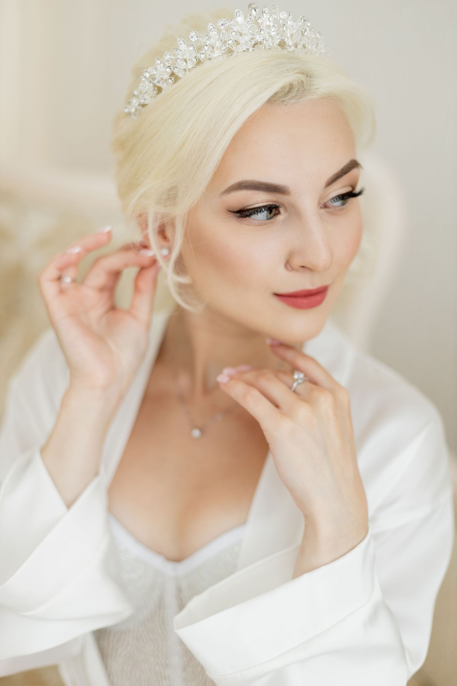 Elegant blonde bride wearing a crystal tiara and white robe, putting on earrings with a diamond ring on her finger.