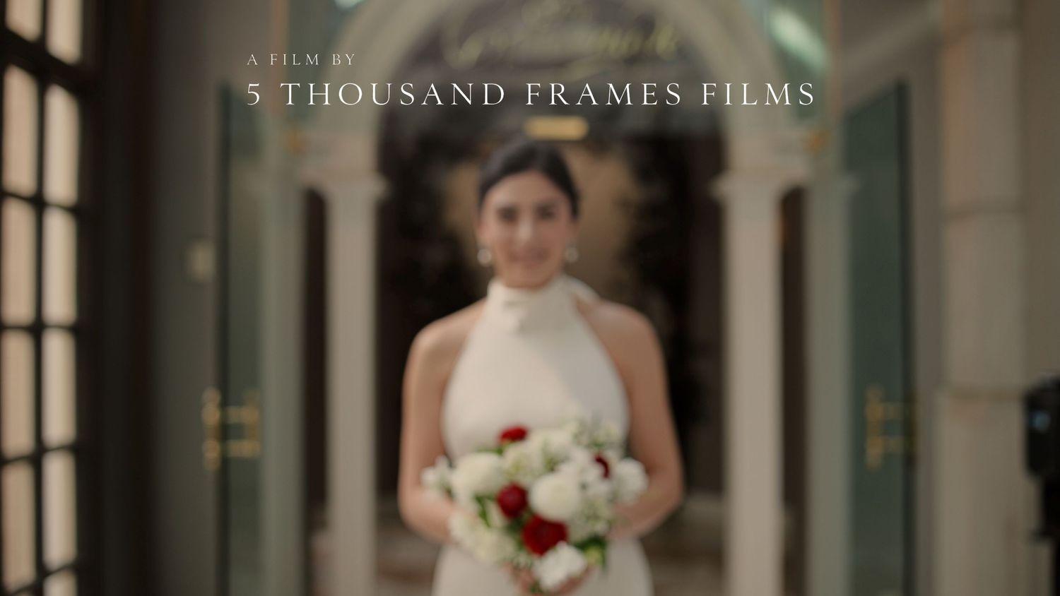 Bride in white halter gown holds red and white bouquet in elegant hallway, film by 5 Thousand Frames Films.