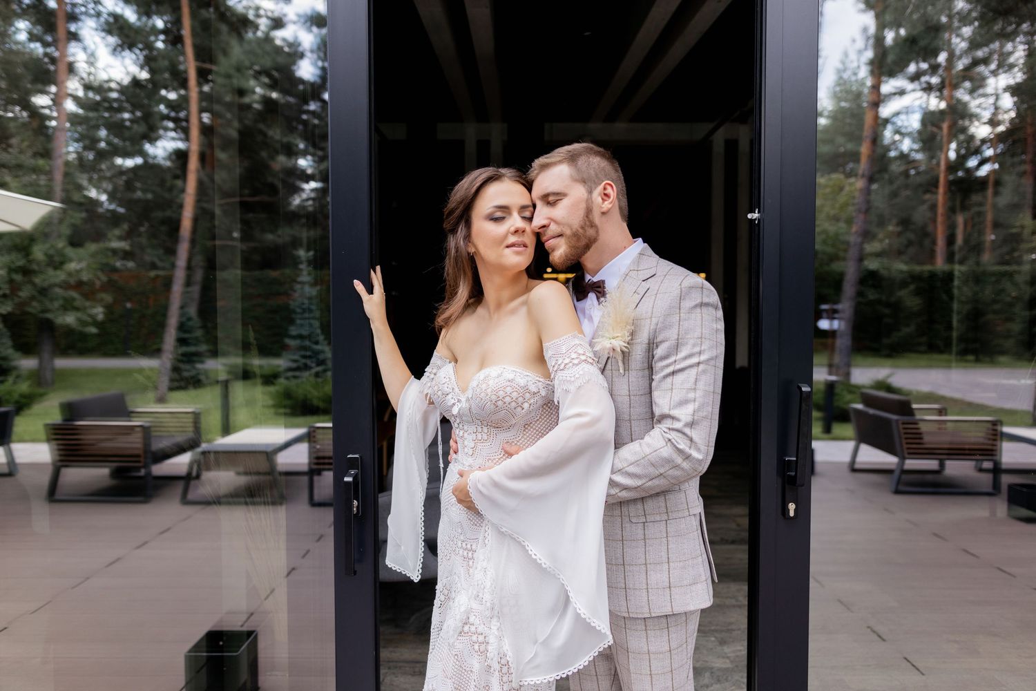 Elegant wedding couple poses for romantic photos against a modern architectural backdrop with pine trees.