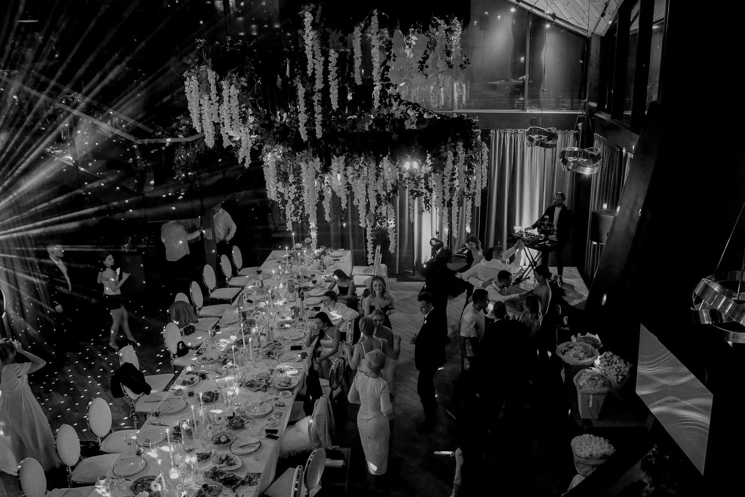 Black and white aerial view of an elegant wedding reception with floral ceiling decor, candlelit tables, and guests dining.