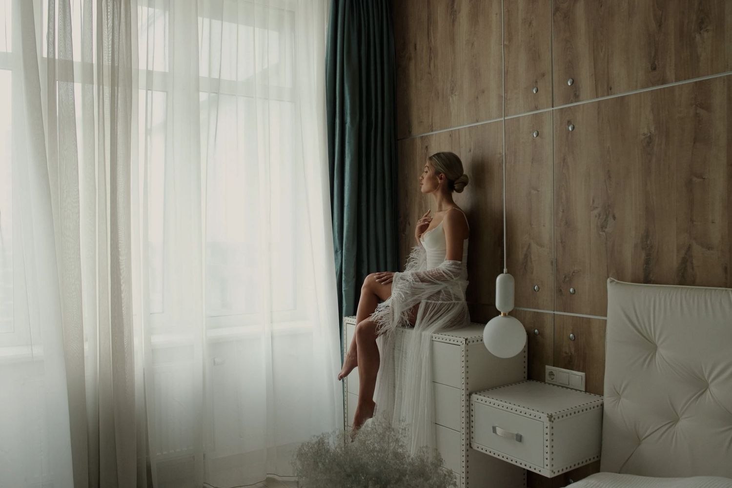 A bride in a white robe sits on a nightstand gazing out a sheer curtained window in an elegant bedroom.