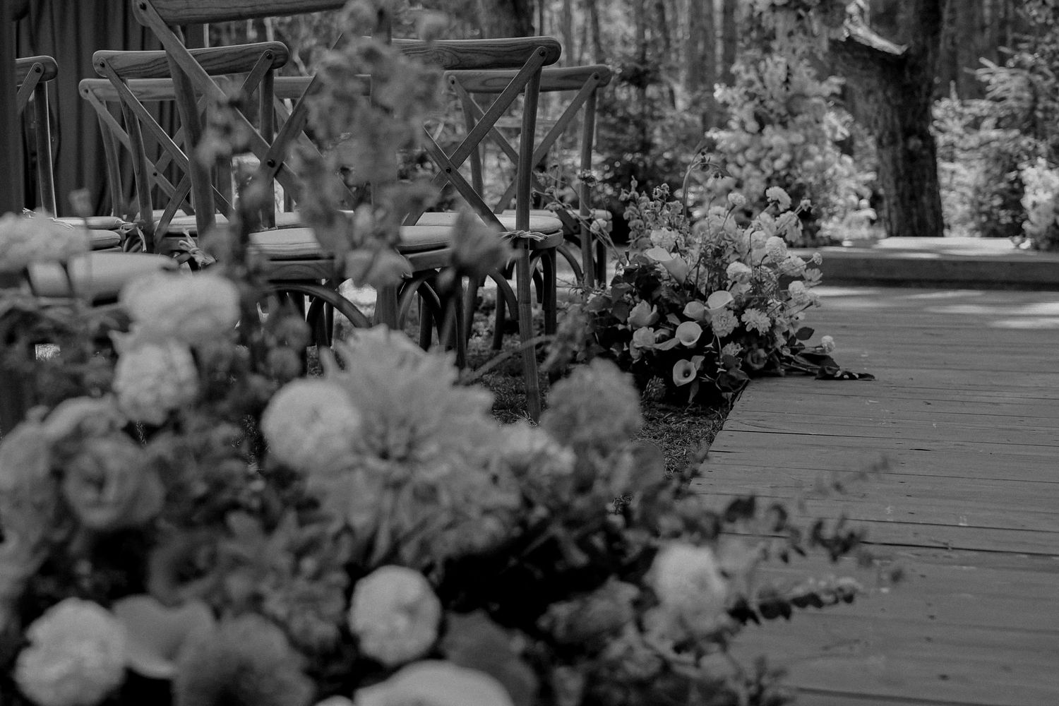 Black and white photo of elegant floral arrangements lining a wooden deck aisle for an outdoor wedding ceremony.