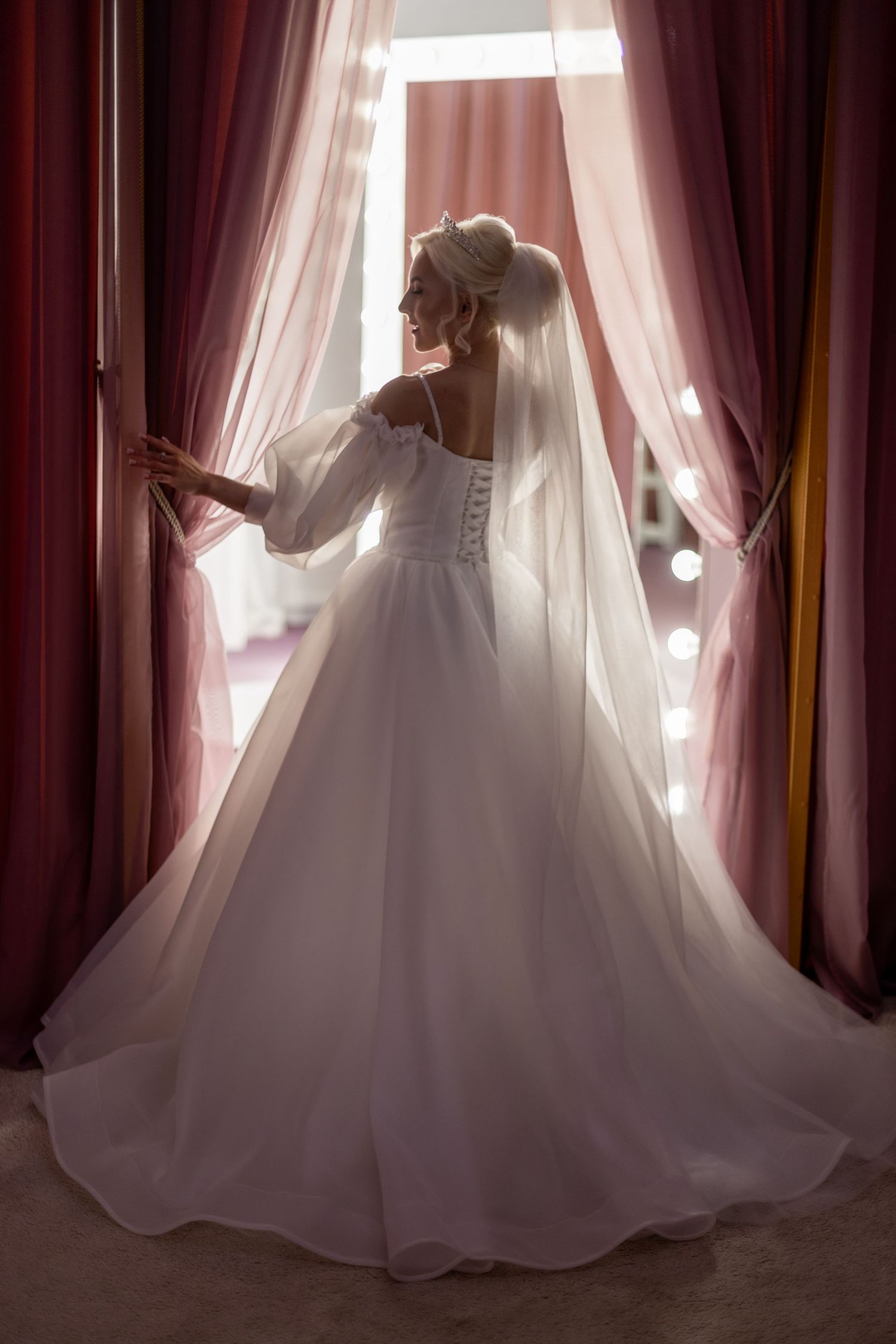 Bride in white ball gown with veil stands at window between pink curtains, viewed from behind.