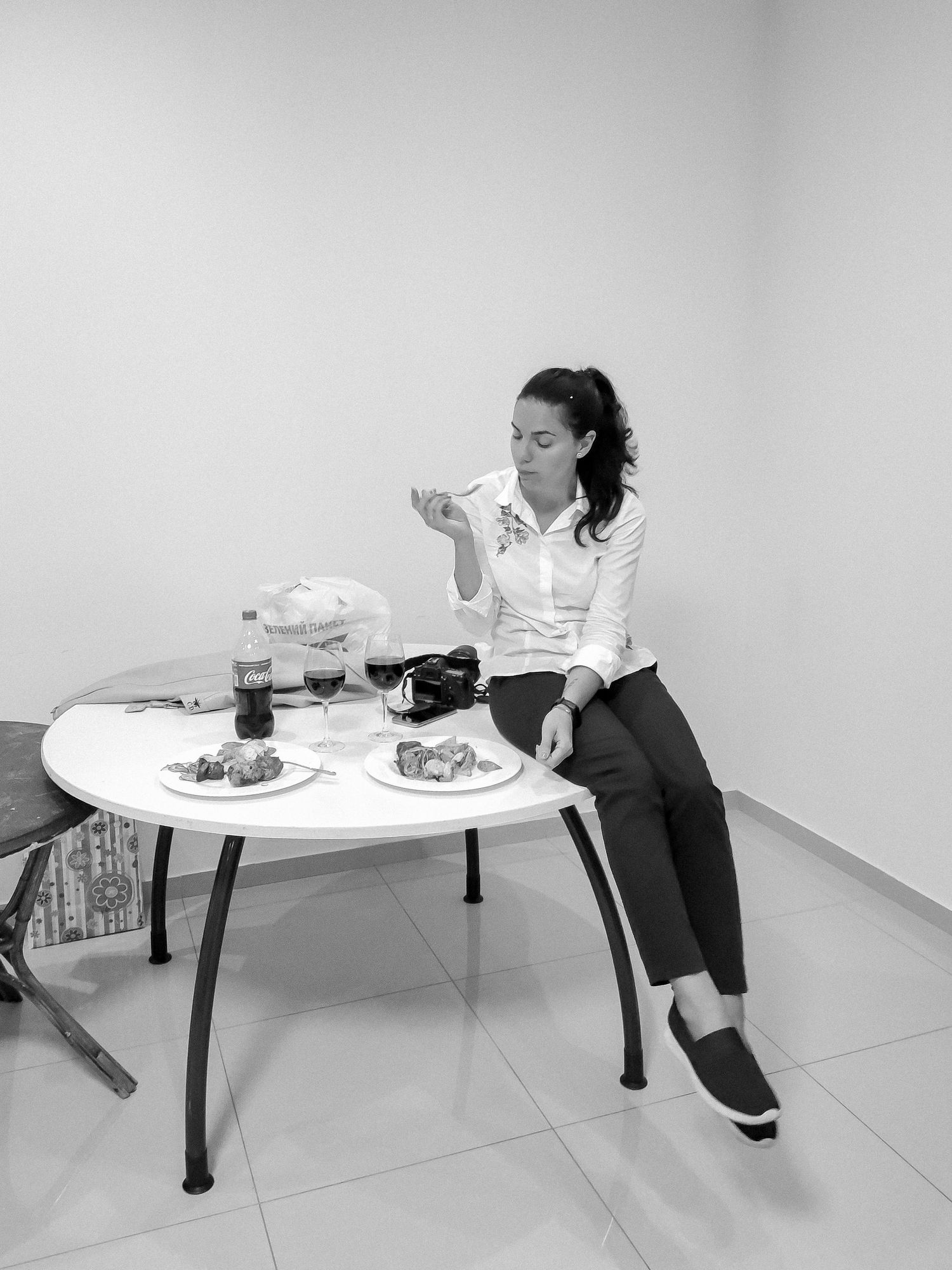 Black and white photo of a woman sitting at a round table eating, with plates of food and a Coca-Cola bottle nearby.